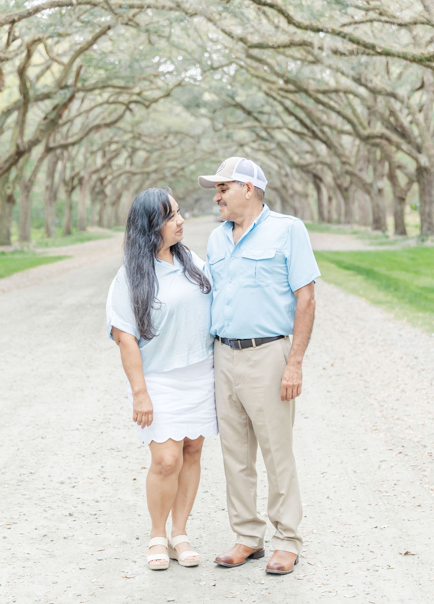 Romantic couple gazing at each other while standing on scenic tree-lined pathway.