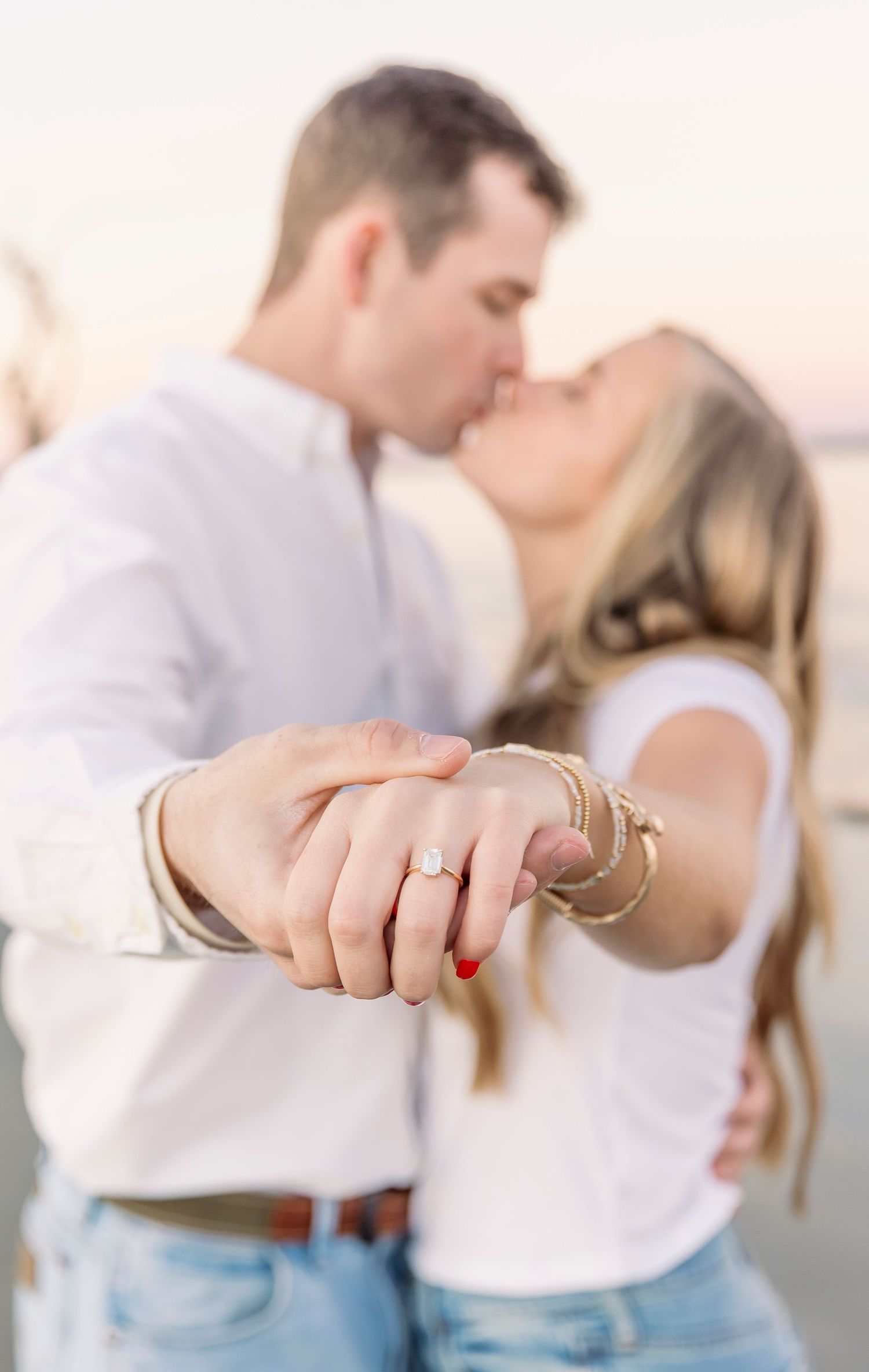 A romantic couple kissing while showcasing an engagement ring in soft golden light.