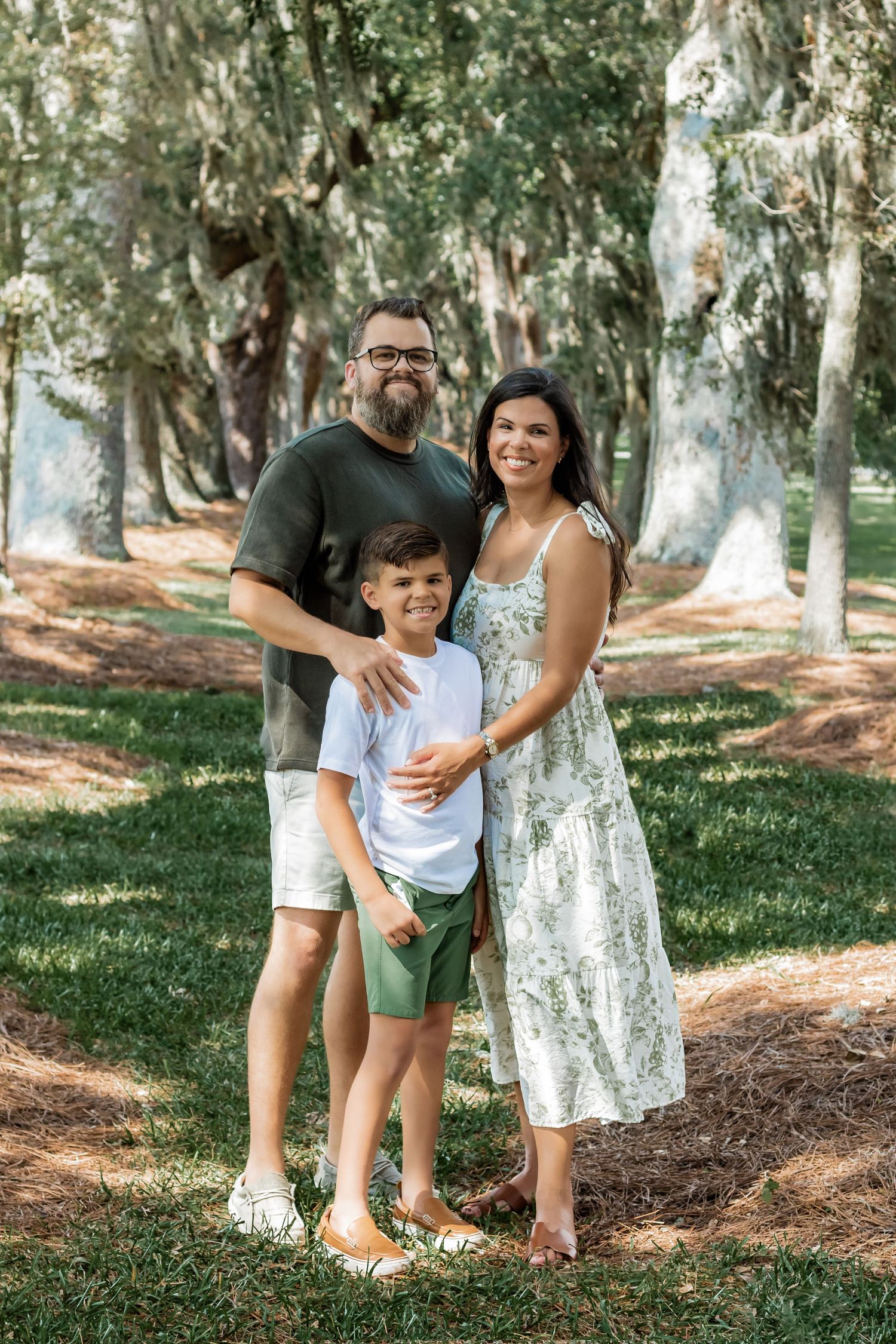 Family of three posing together outdoors among Spanish moss-covered trees on a sunny day.
