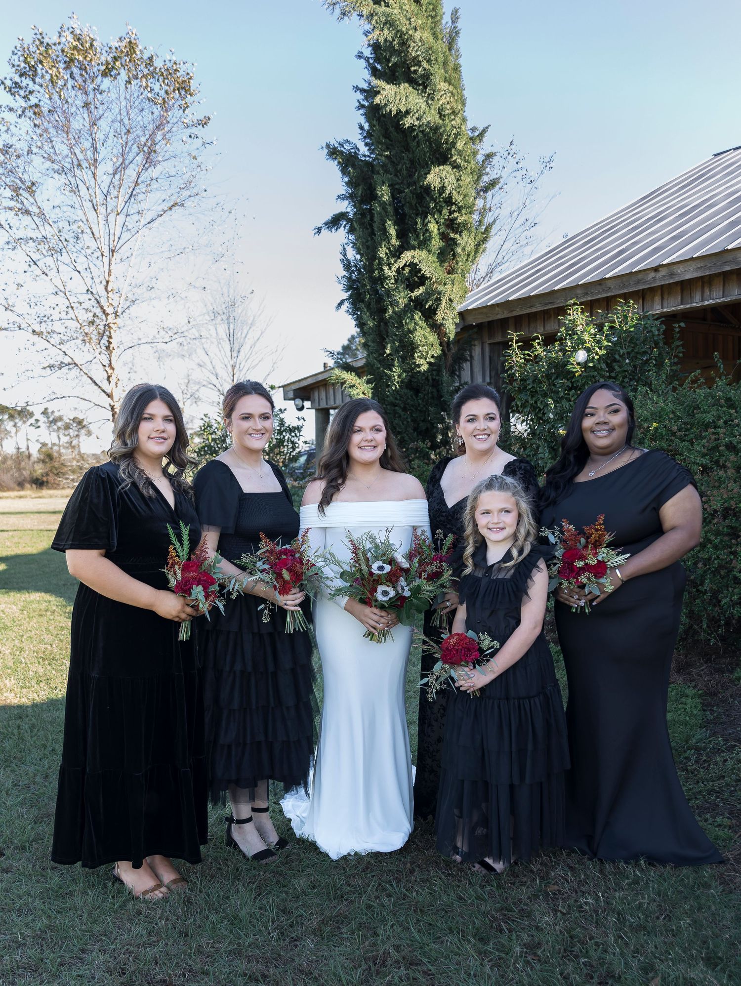 Wedding party in black dresses poses with bride in white, holding burgundy bouquets outdoors.