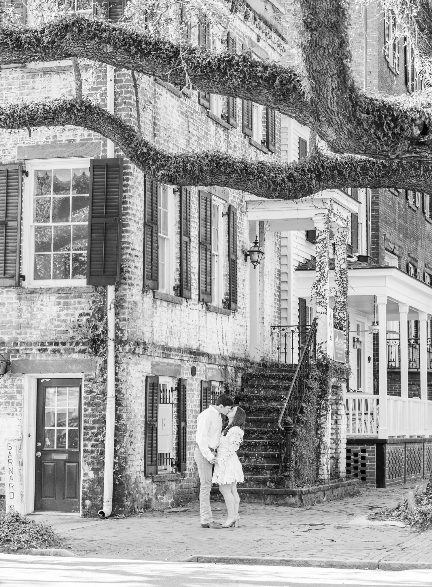 A black and white image of a couple embracing in front of a historic brick building.