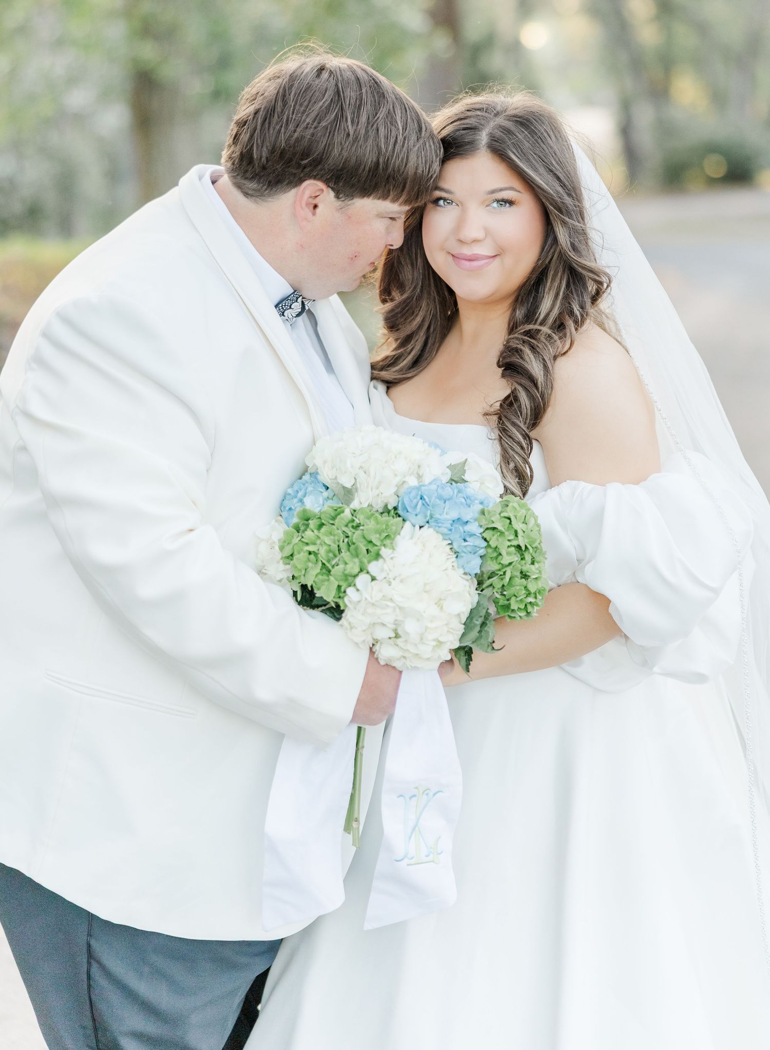 Bride and groom in white attire share a tender moment, bride holding blue and white hydrangea bouquet outdoors.