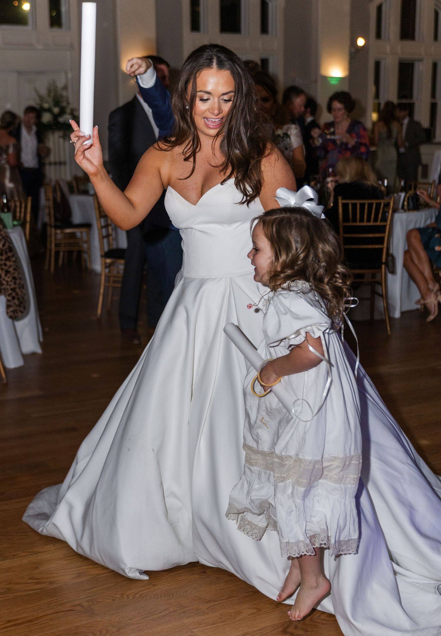 Bride in white gown dances with young flower girl at wedding reception.