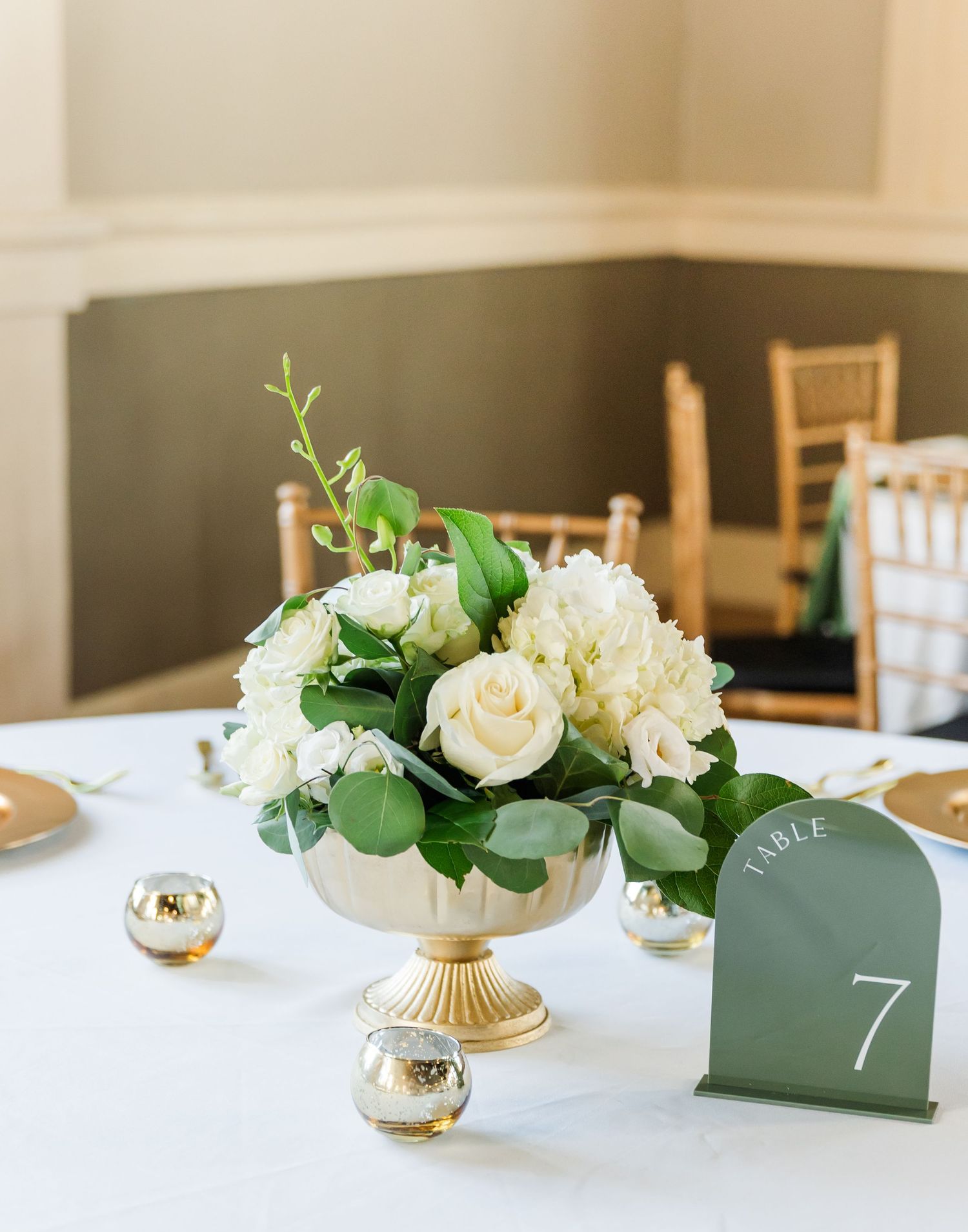 Gold pedestal vase with white roses and greenery centerpiece beside a sage green Table 7 number card.