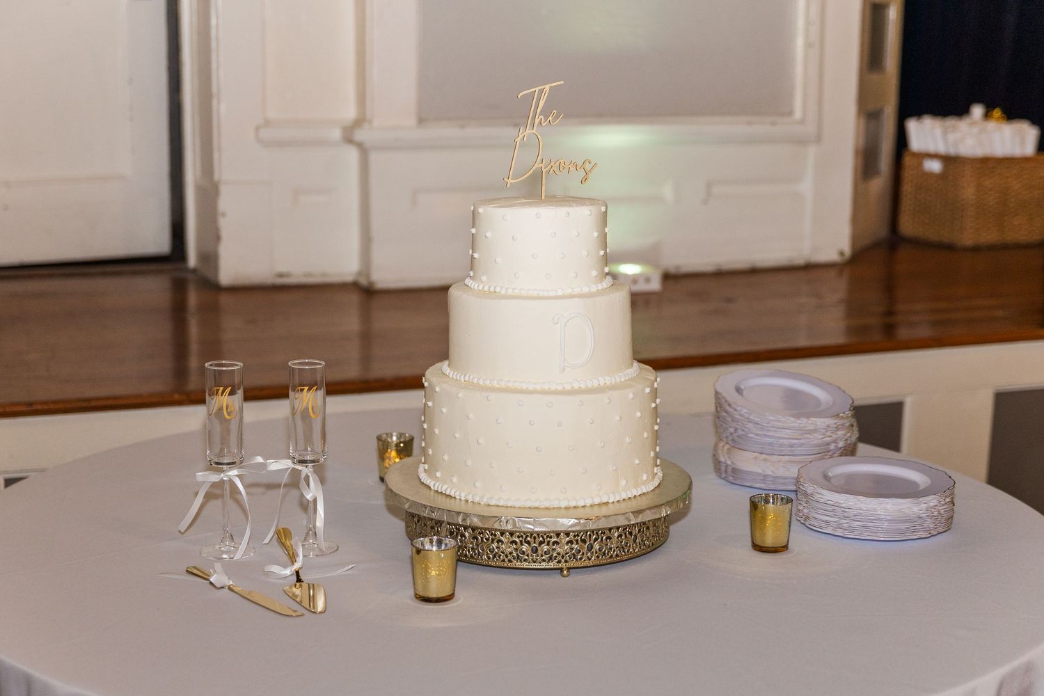 Elegant four-tier white wedding cake on gold stand with champagne flutes and candles on a round table.