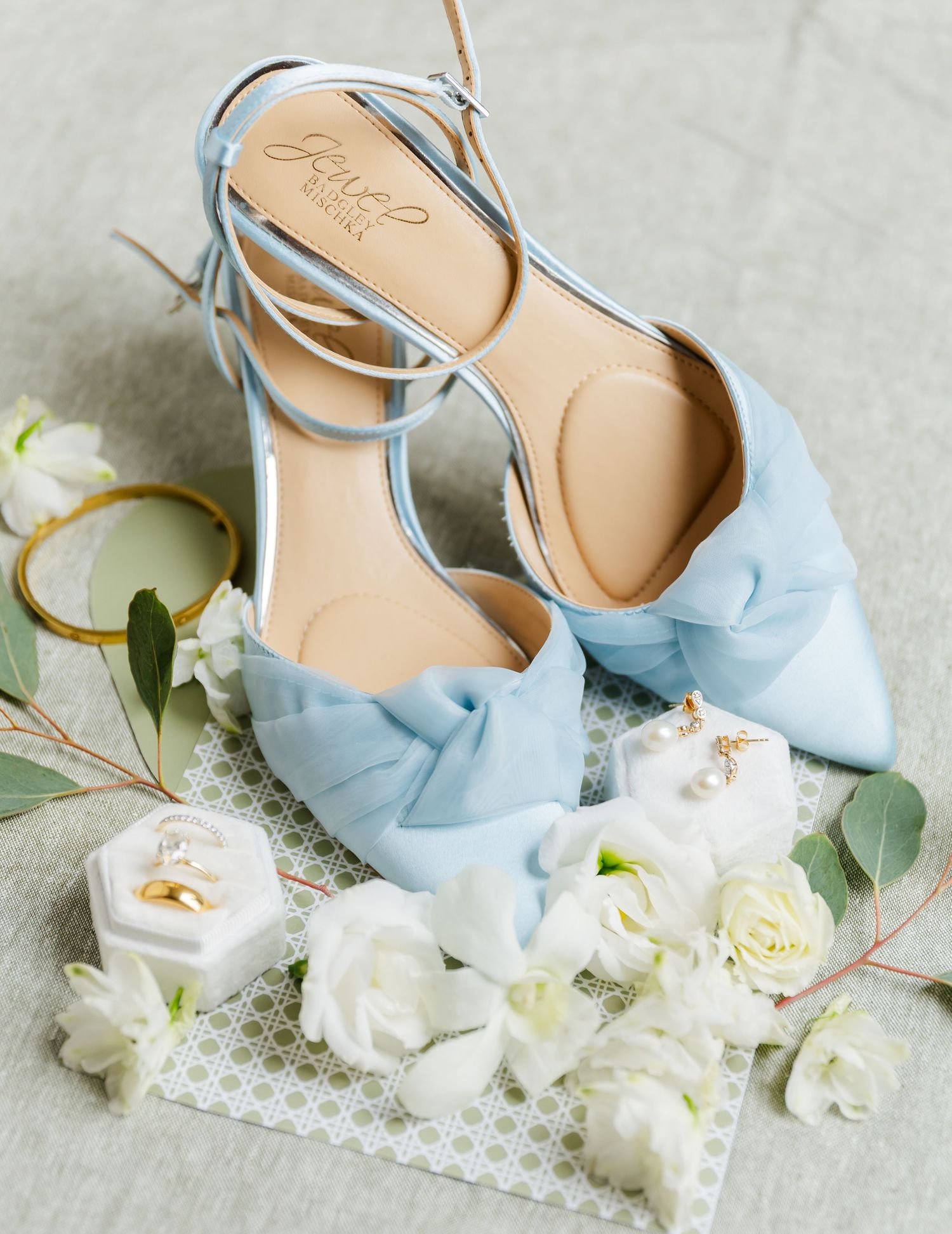 Light blue satin bow heels by Joie surrounded by white roses, wedding rings, and eucalyptus.