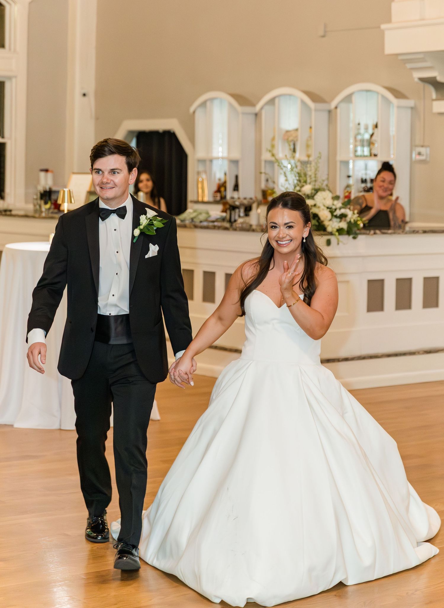 Bride in white ball gown and groom in black tuxedo hold hands while making their grand entrance at wedding reception.