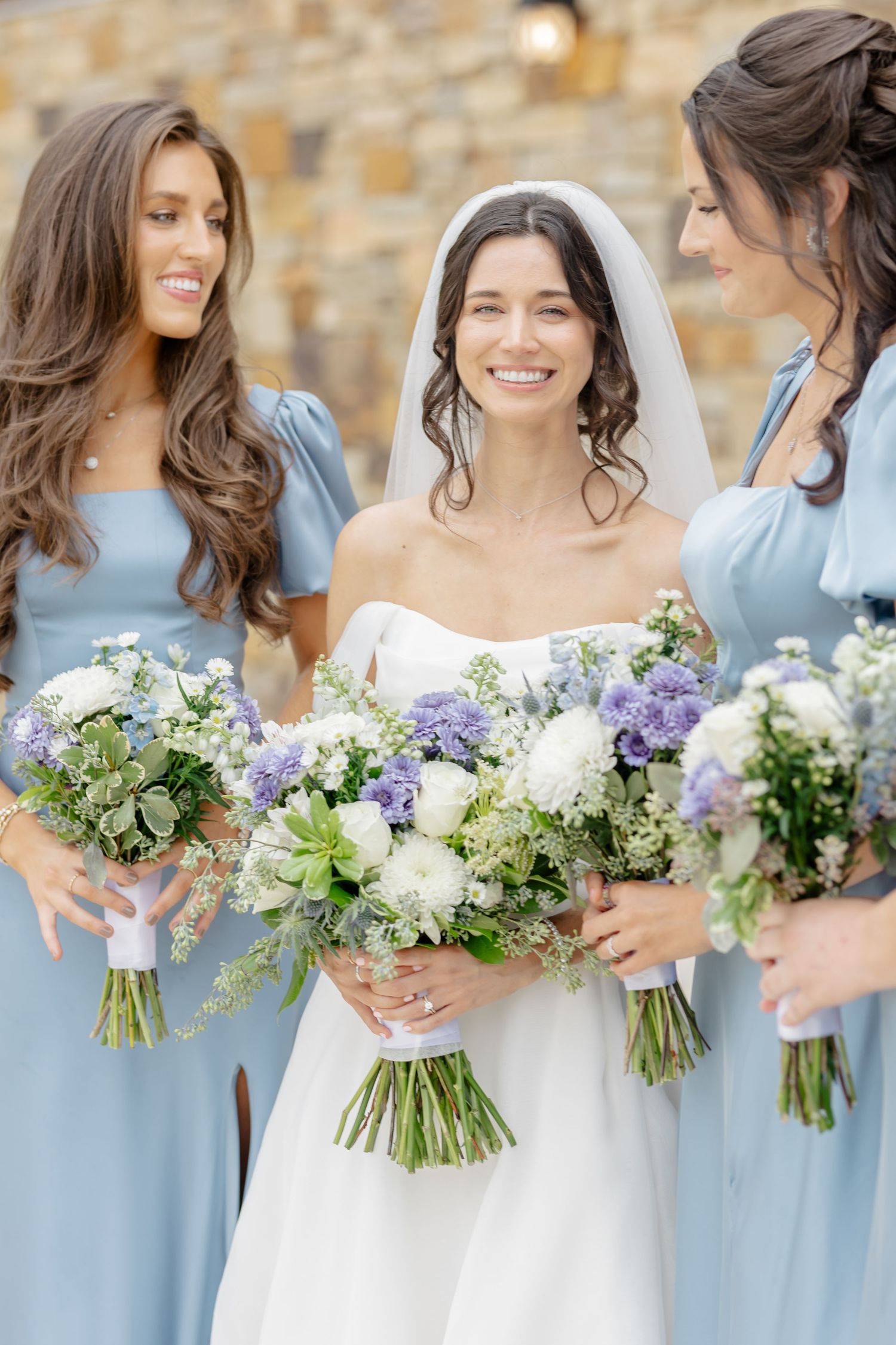 A row of bridesmaids in light blue dresses holding elegant bouquets of white, purple and green flowers against a stone wall.
