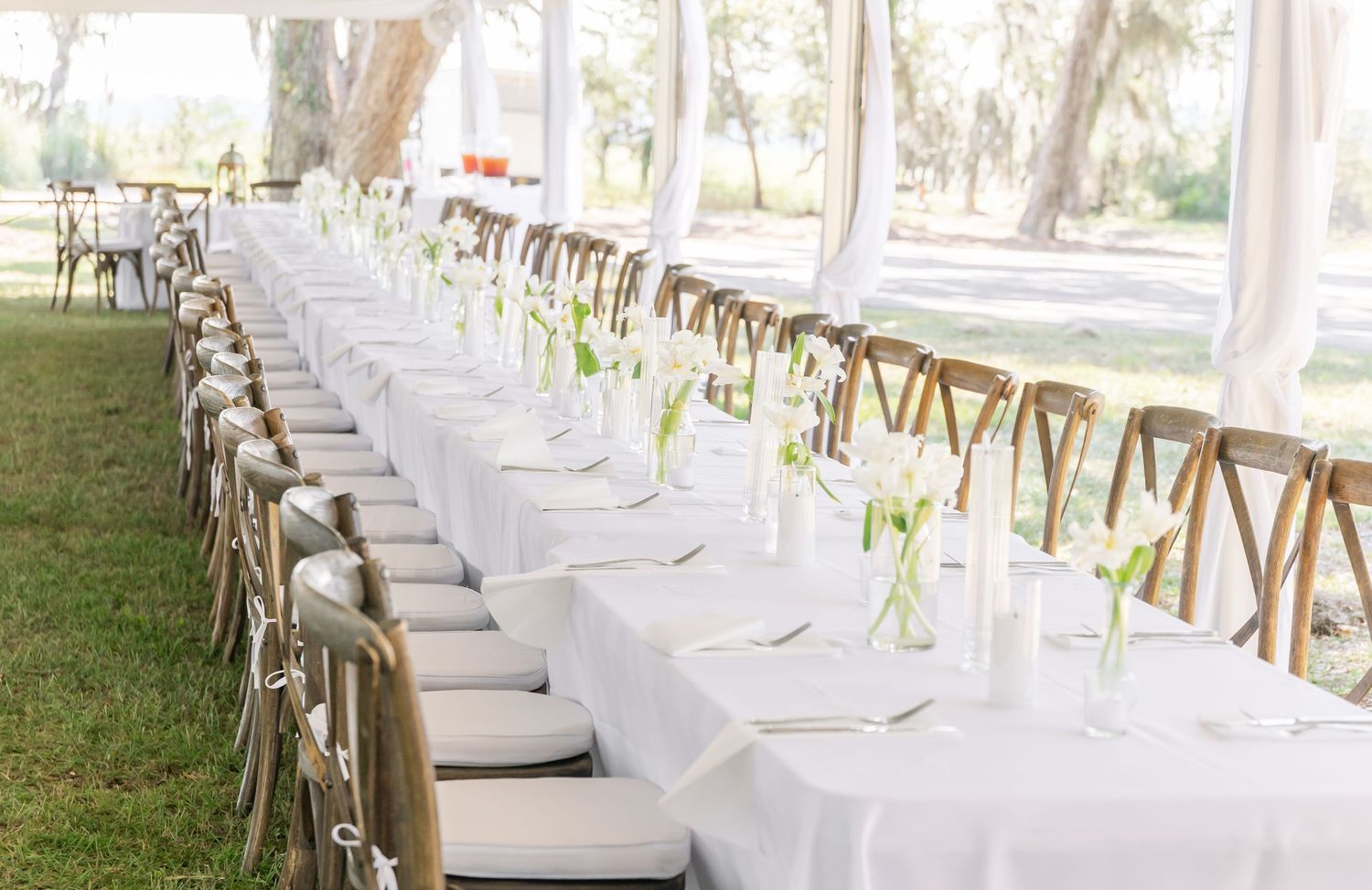 Long white dining tables with wooden cross-back chairs set up under trees for an outdoor wedding reception.