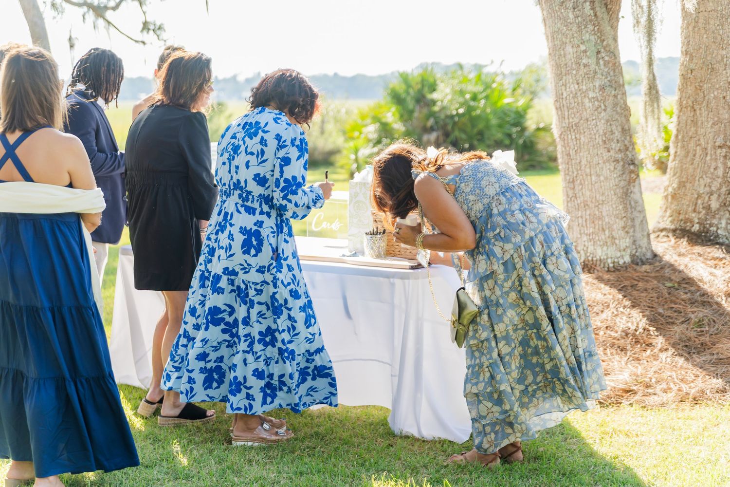 A group of people in blue dresses gather outdoors near tables under trees at a summer event.