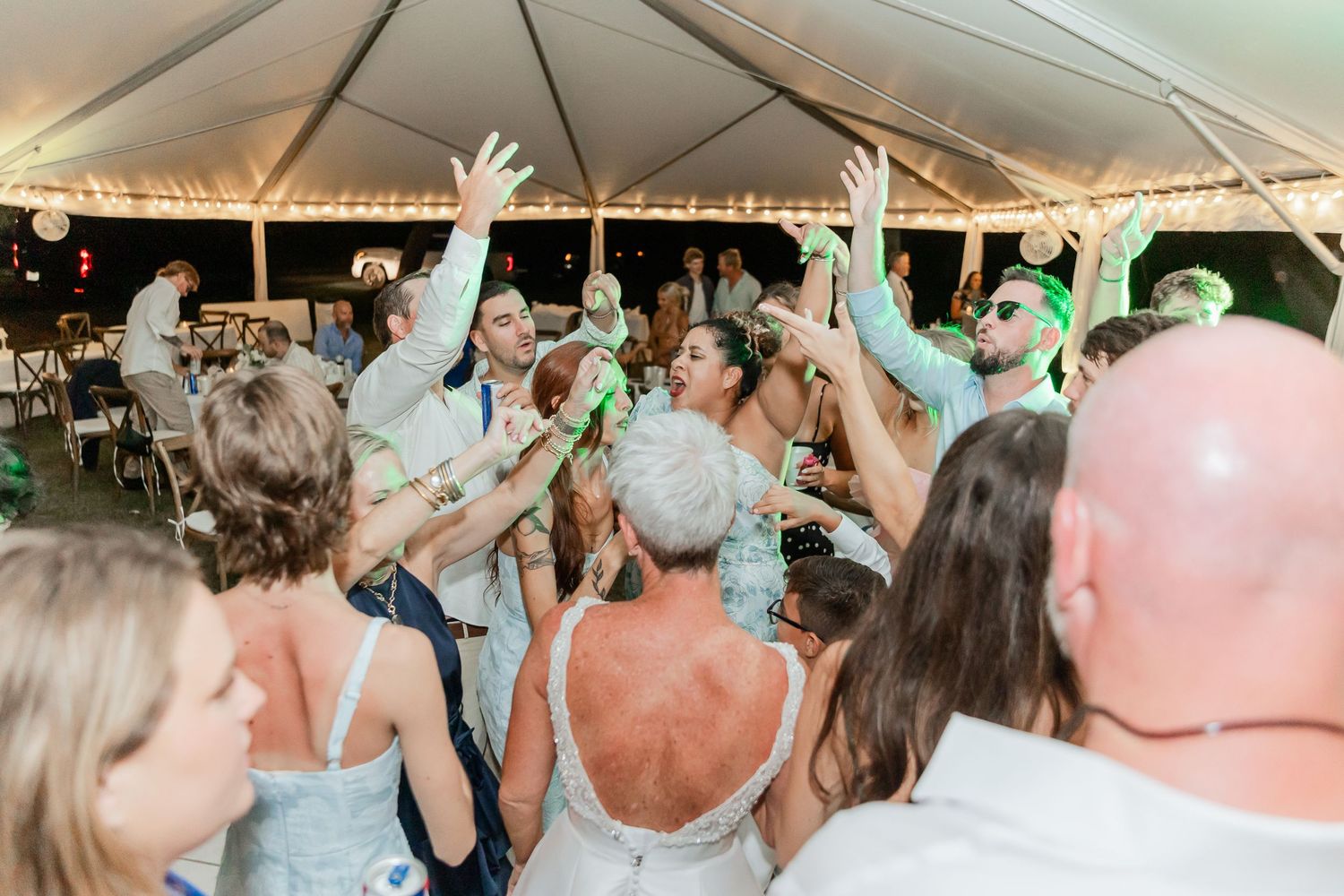 Wedding reception guests dancing and celebrating under a white tent with string lights overhead.