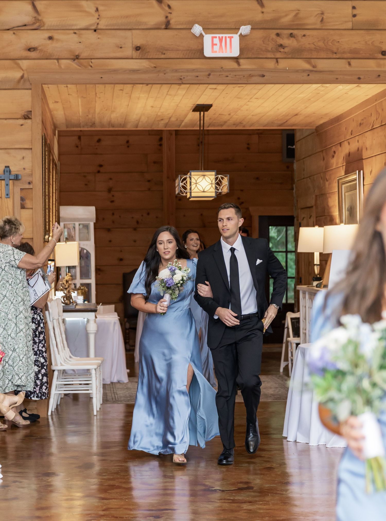 Wedding guests mingle and dance in a rustic wooden reception venue with exposed beams and warm lighting.