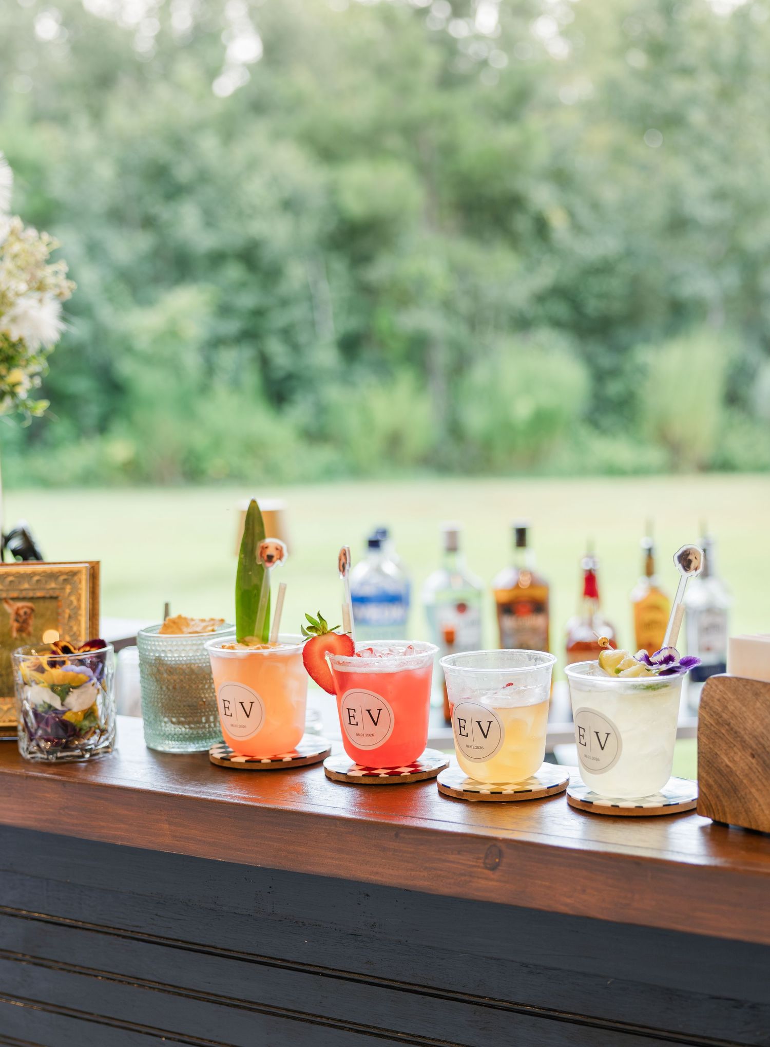Colorful monogrammed wedding cocktails lined up on wooden bar with liquor bottles and garden backdrop.