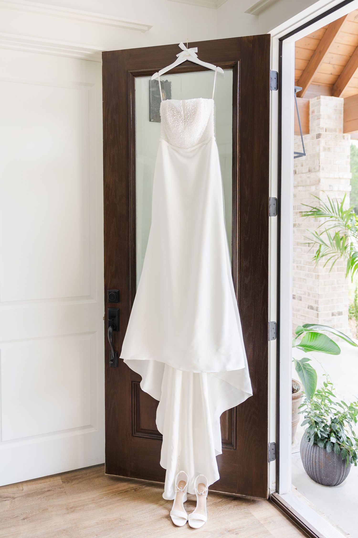 Elegant strapless white wedding dress hanging on a dark wood door with white bridal heels below.