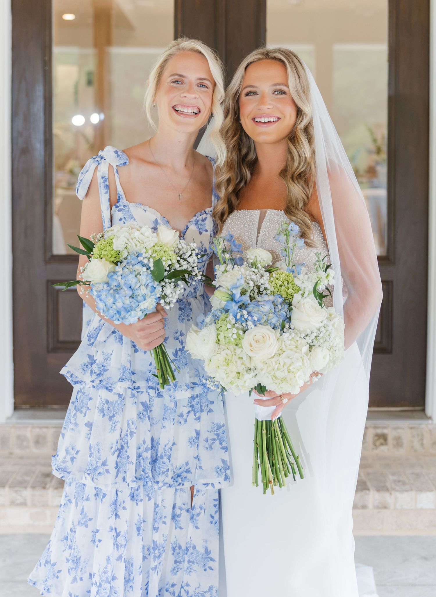 Bride and bridesmaid smiling together holding matching white and blue hydrangea bouquets at wedding.