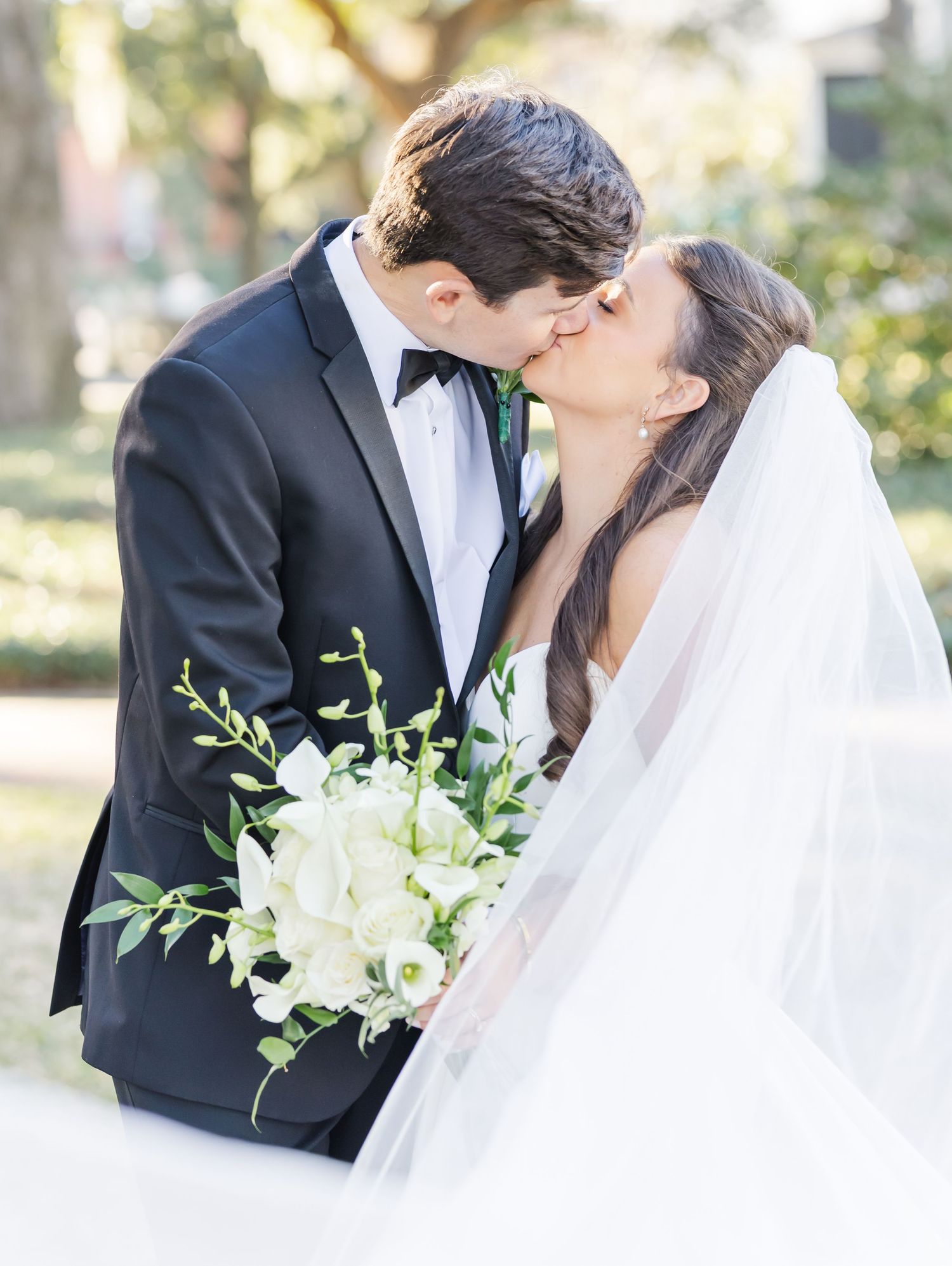 A romantic wedding moment with a couple sharing a kiss while holding a white floral bouquet in an outdoor garden setting.