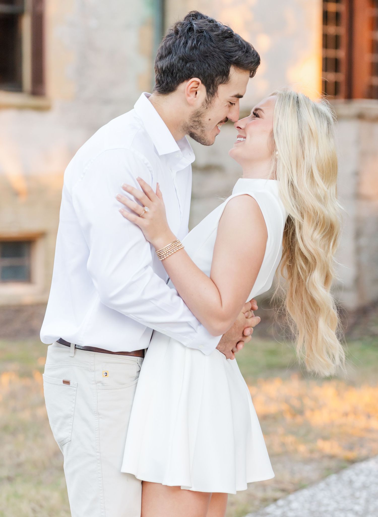 A romantic couple in white outfits embraces during a sunset photoshoot in a rustic outdoor setting.