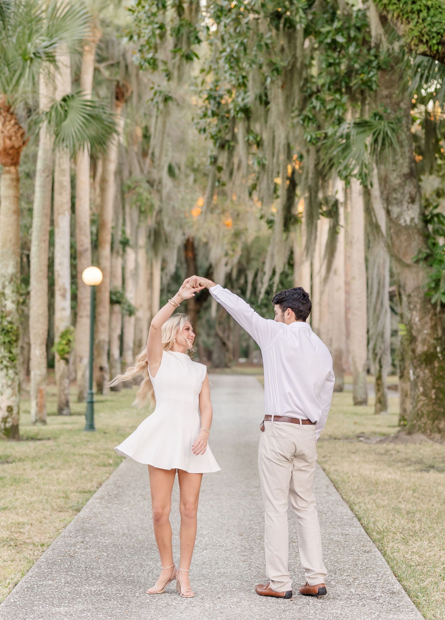 A romantic couple in white and beige outfits poses playfully on a tree-lined path with Spanish moss.