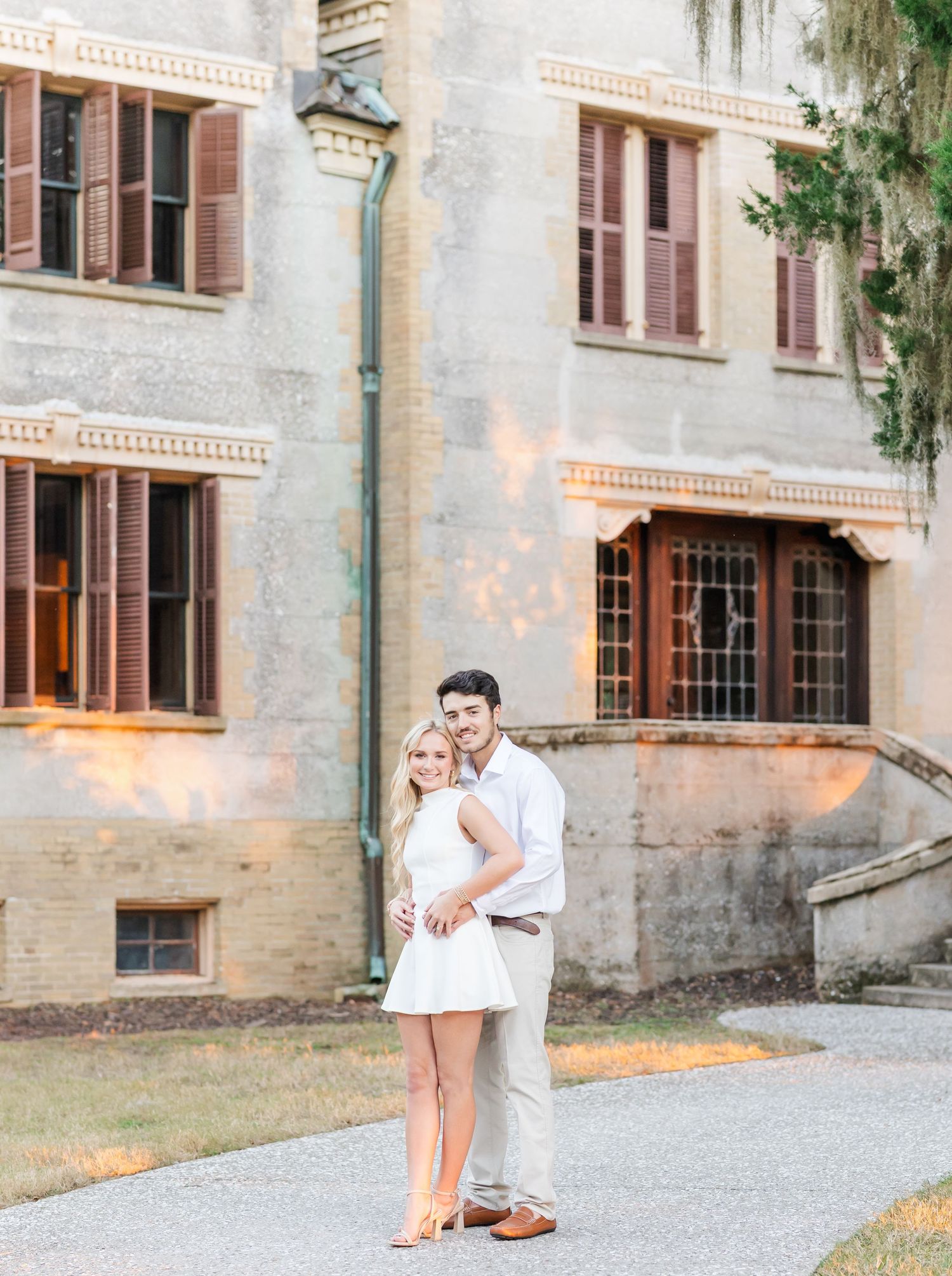 A couple shares a romantic moment on a gravel path outside a historic stone building at sunset.