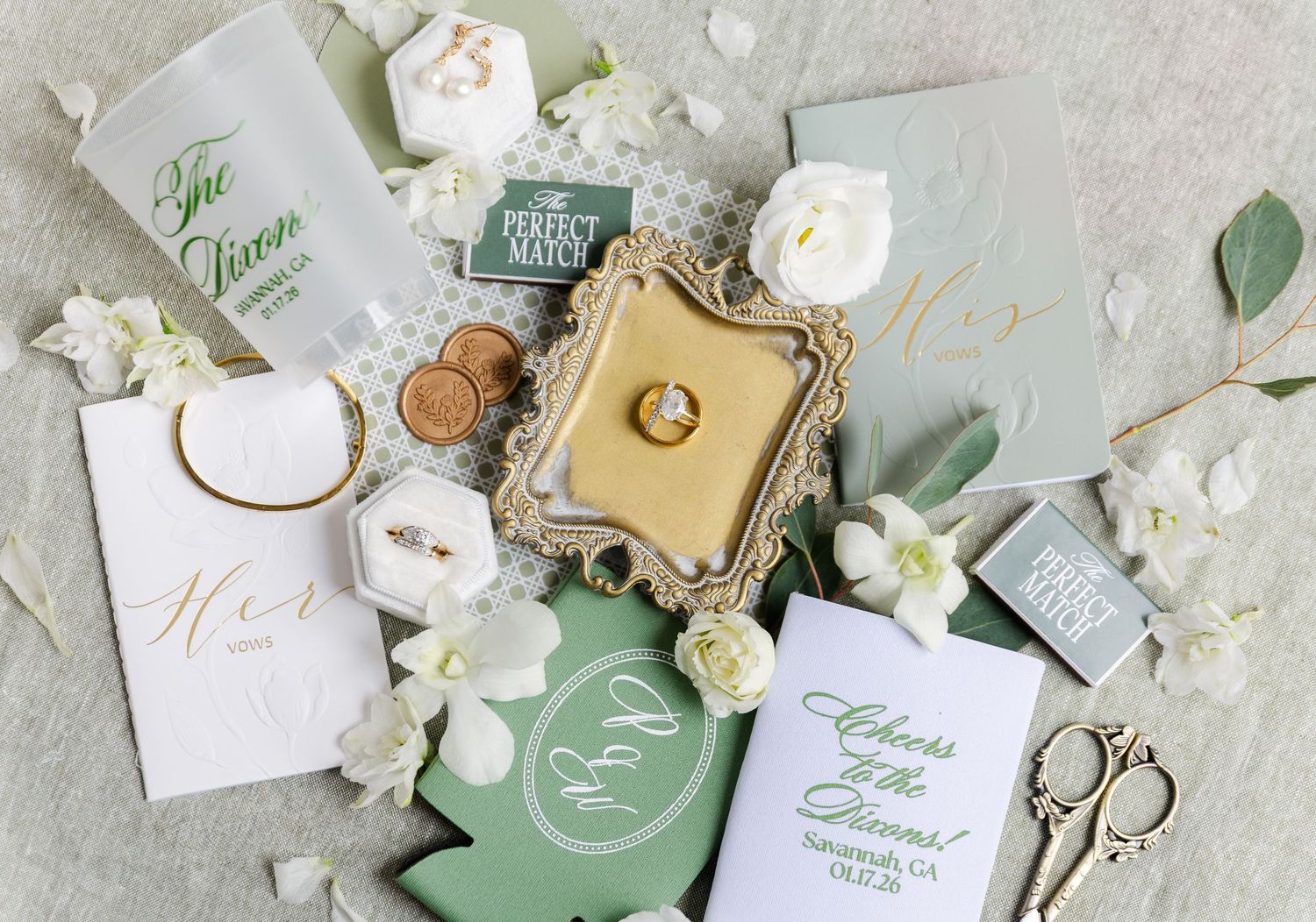 Wedding invitation suite with white flowers, gold tray, rings, and sage green accents arranged on a light gray surface.