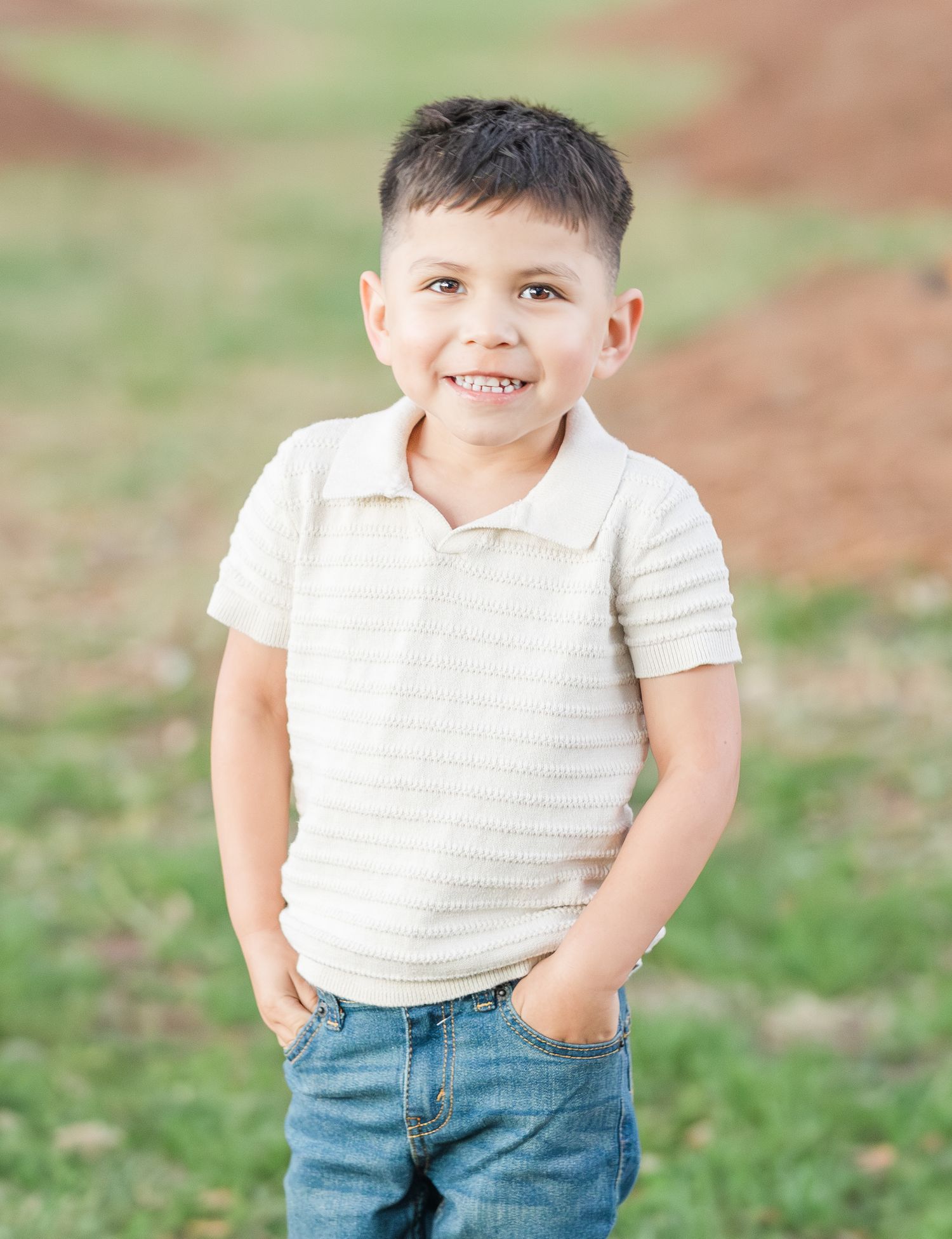 Young child in striped polo shirt smiles brightly while posing outdoors with hands in pockets.