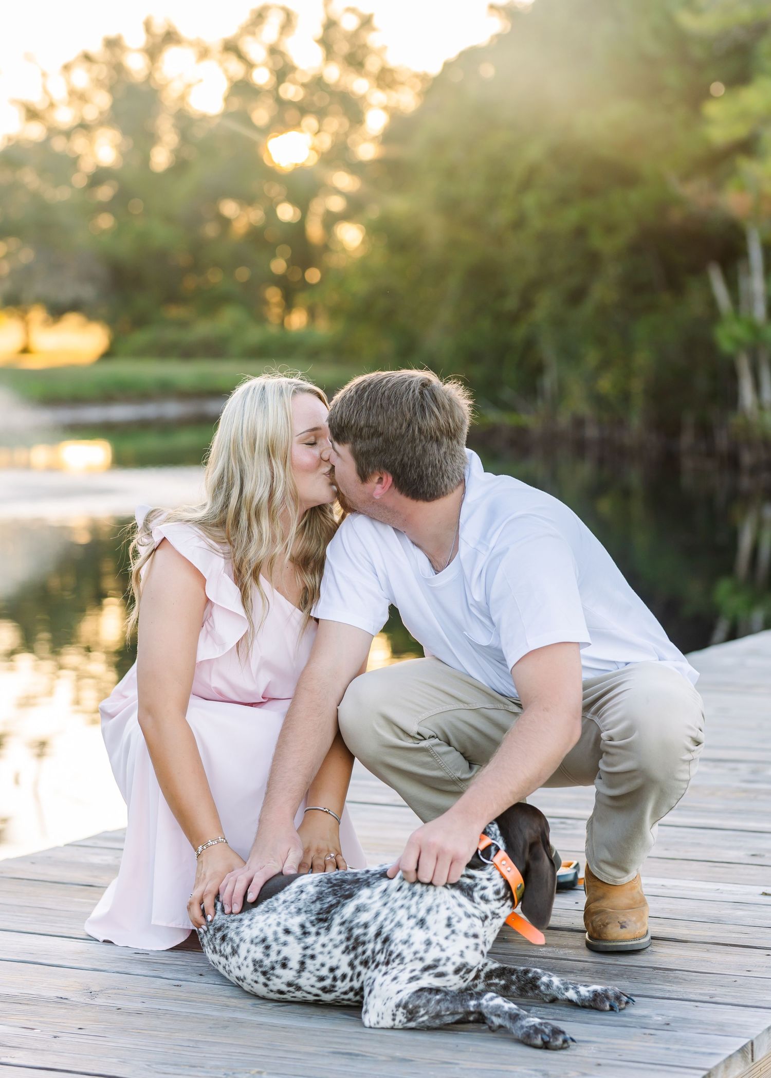 Couple shares tender moment with their spotted dog on wooden dock at sunset.
