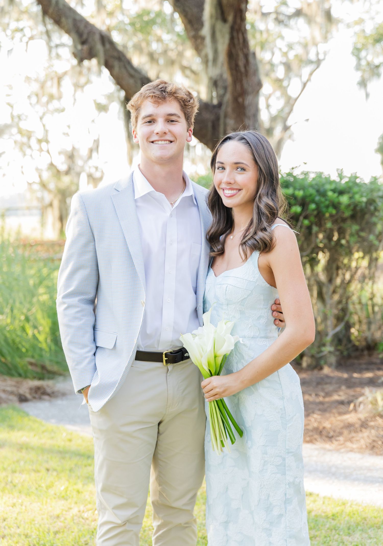 A couple in wedding attire poses outdoors against a natural backdrop with trees and sunlight filtering through.