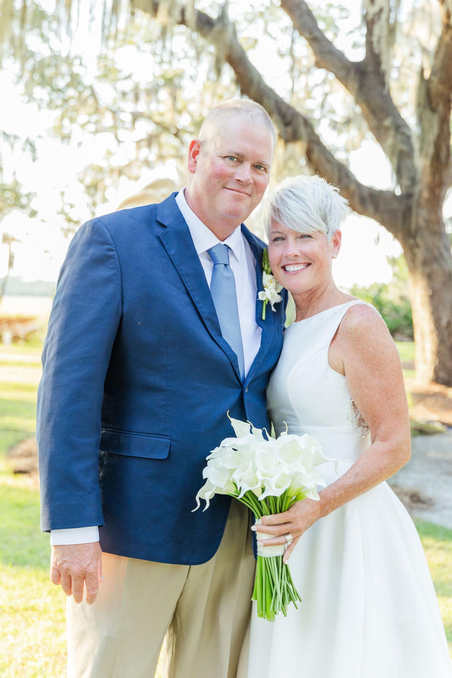 A wedding couple in navy blue and white attire share a joyful moment outdoors under trees.