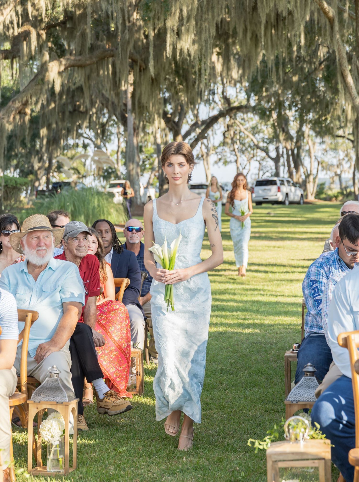 Outdoor wedding ceremony with guests seated on wooden chairs under moss-draped trees in a grassy setting.