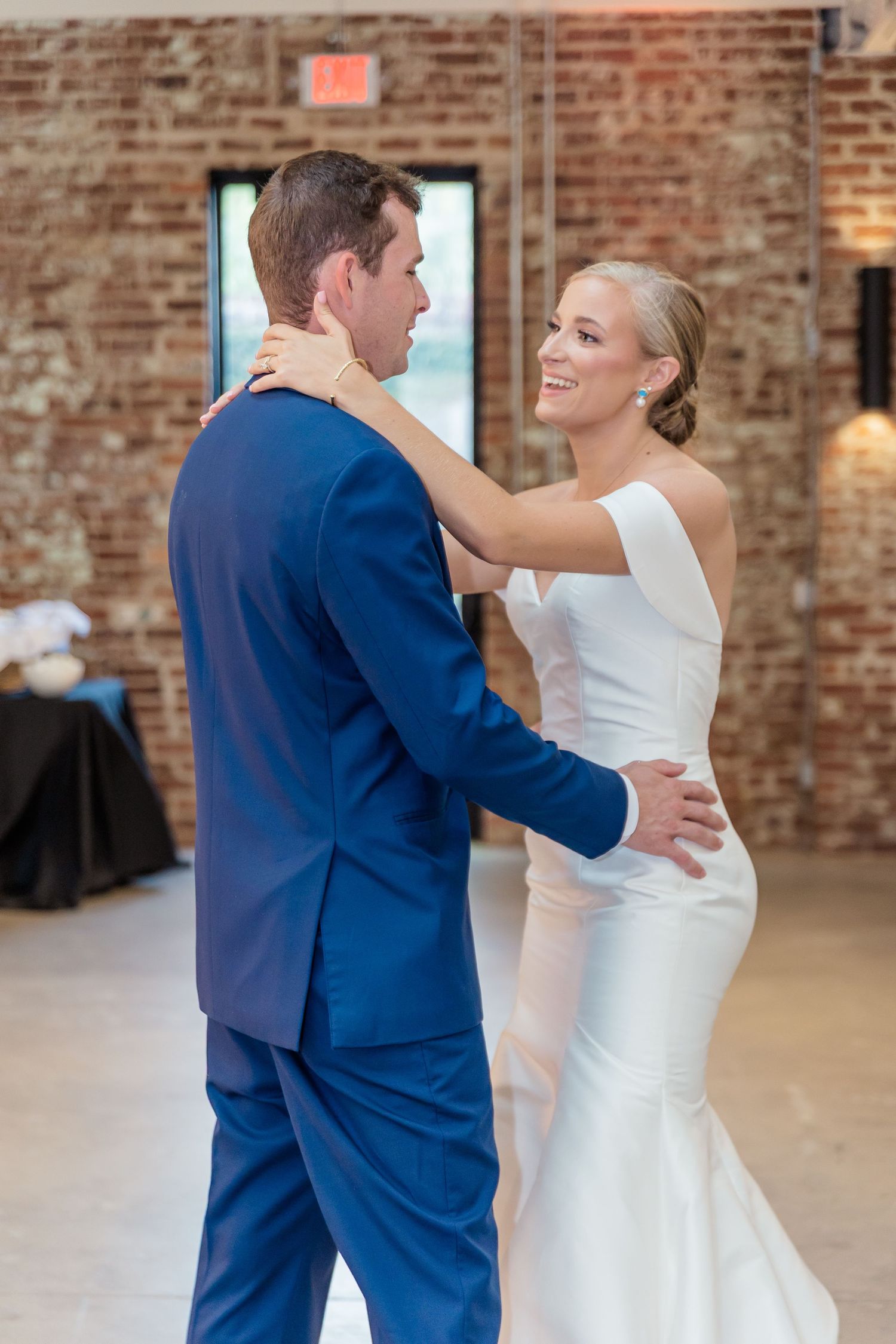 A couple in navy blue and white formal attire share a romantic dance together in a rustic brick-walled venue.