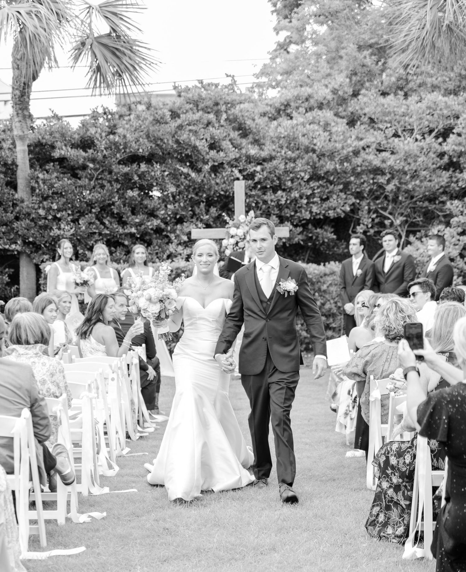 Black and white panoramic photo of an outdoor wedding ceremony with white chairs arranged on a lawn under palm trees.