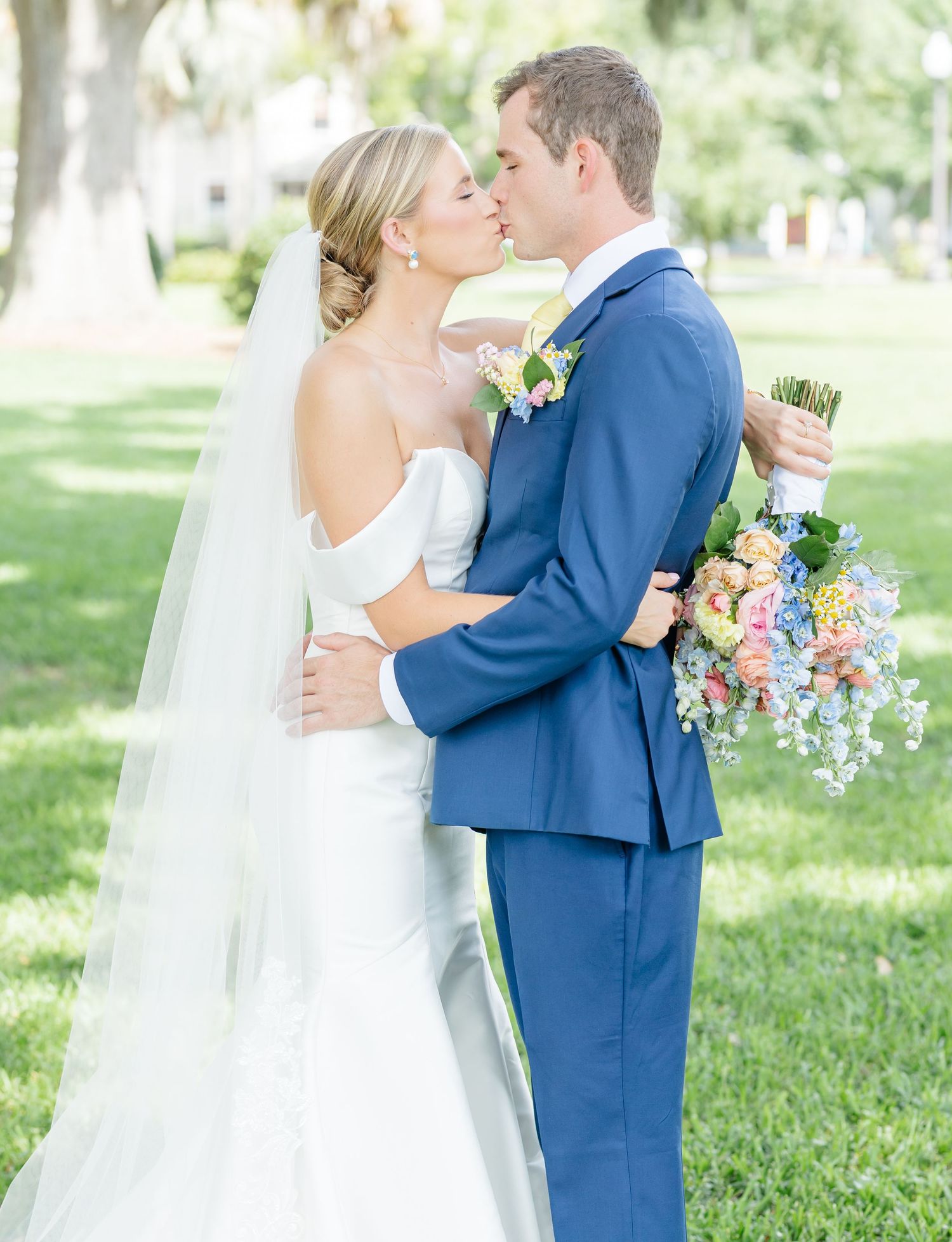 A couple shares a romantic kiss in a garden setting while wearing wedding attire, the bride in white and groom in navy blue.
