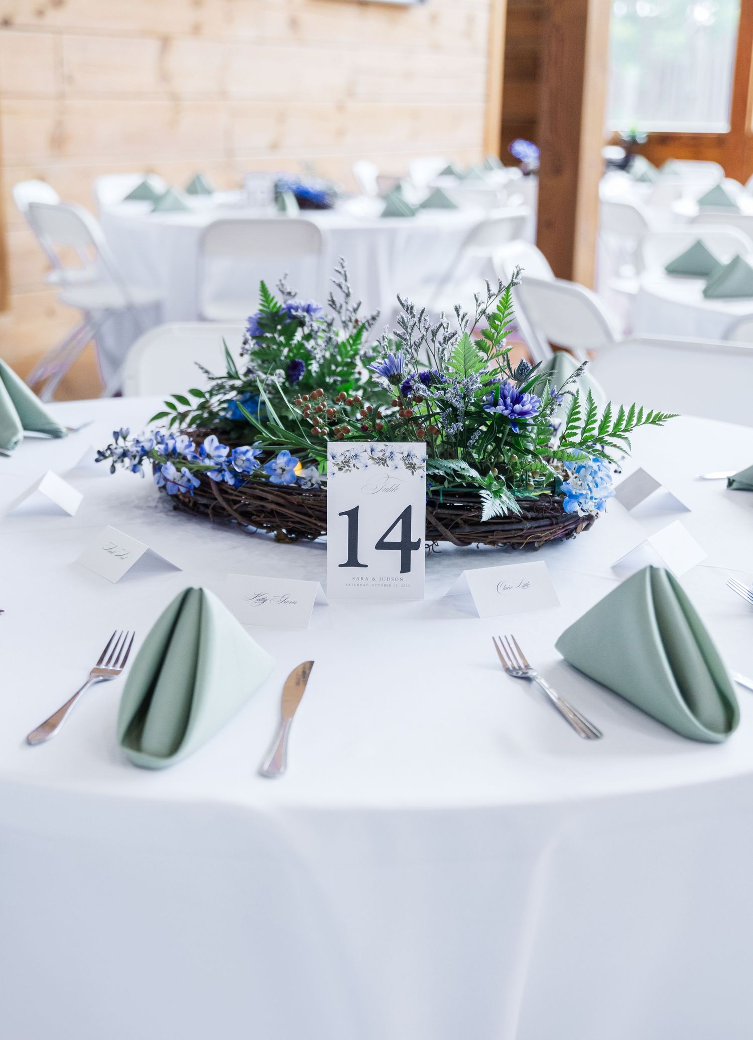 Wedding table setting with sage green napkins and rustic floral centerpiece featuring blue flowers and table number 14.