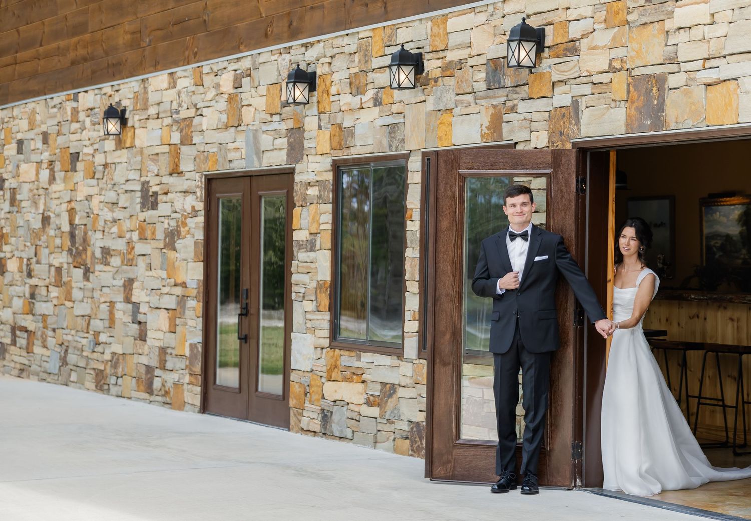 A couple in formal wedding attire poses together against a rustic stone wall exterior of a building.