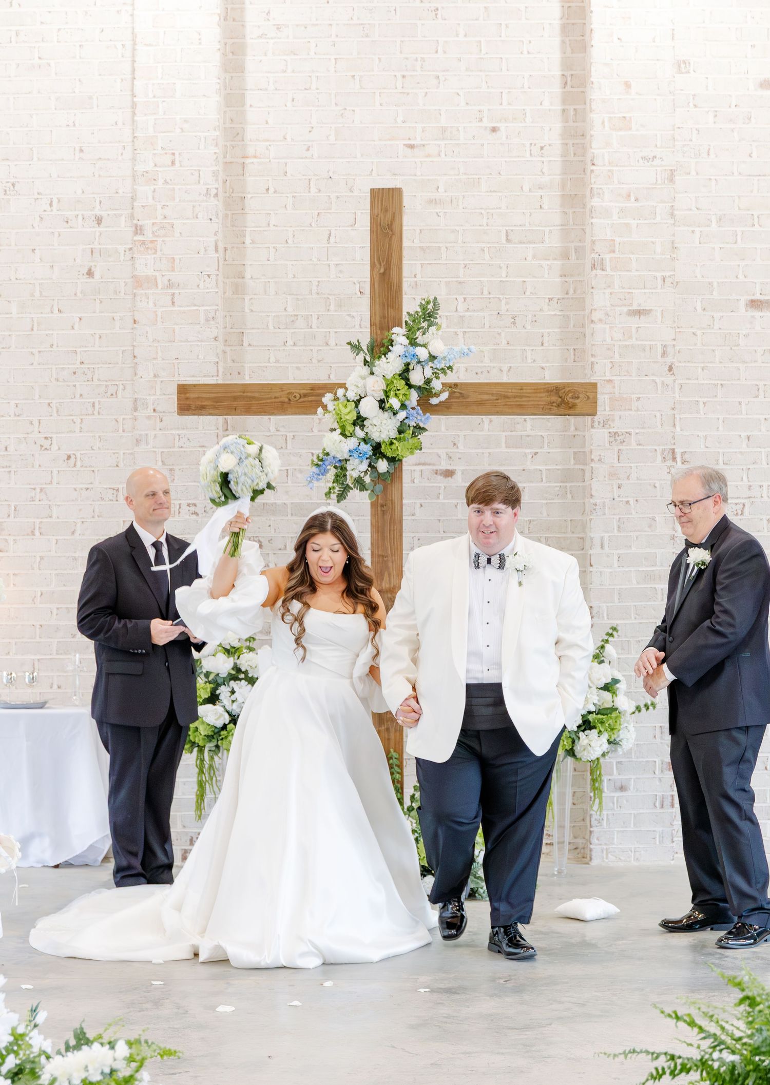 Bride and groom holding hands at the altar beneath a wooden cross adorned with blue and white floral arrangements.