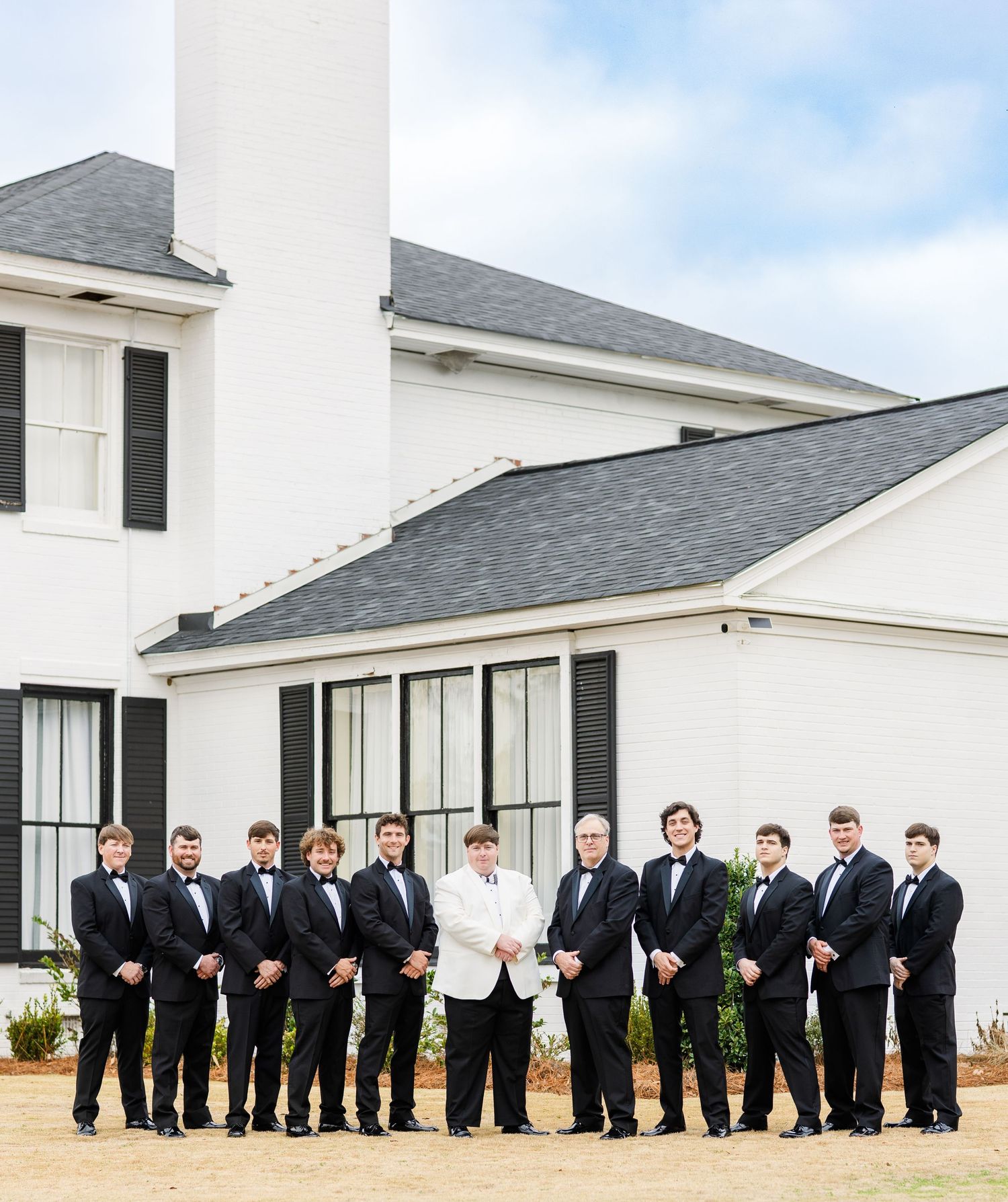 Groom in white jacket with groomsmen in black tuxedos posing outside a white modern venue.