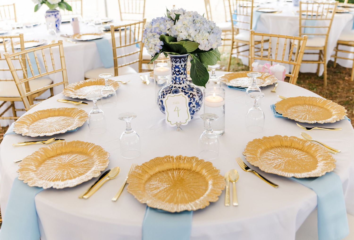 Elegant wedding reception table with gold charger plates, blue hydrangea centerpiece, and light blue table runner.