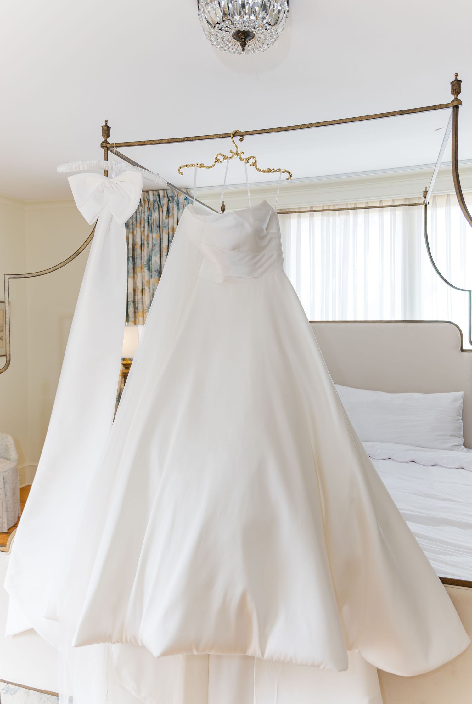 Elegant strapless white ball gown wedding dress hanging on a gold canopy bed frame in a bright bridal suite.