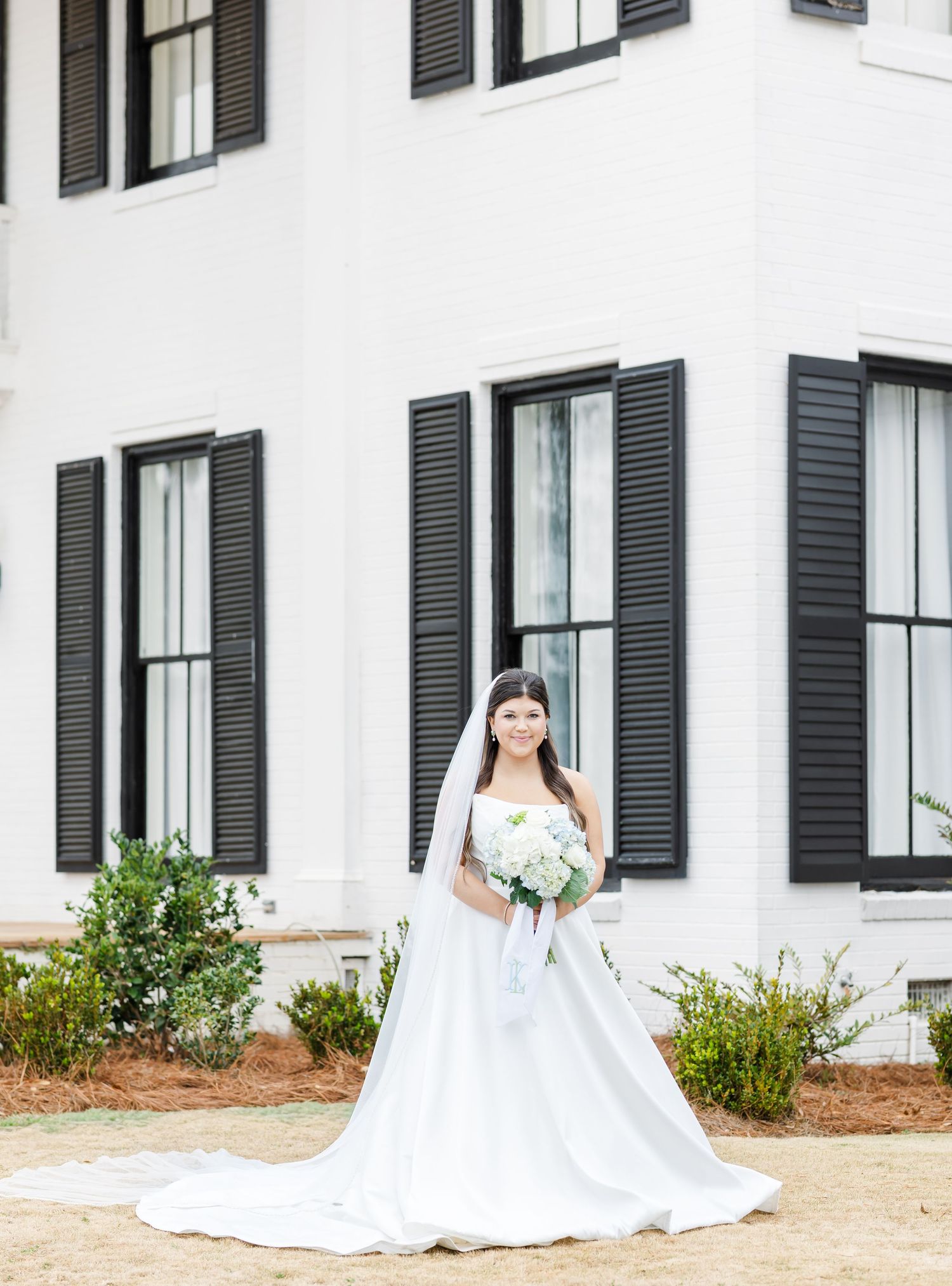 Bride in strapless white ball gown with veil and bouquet poses outside white colonial-style venue with black shutters.