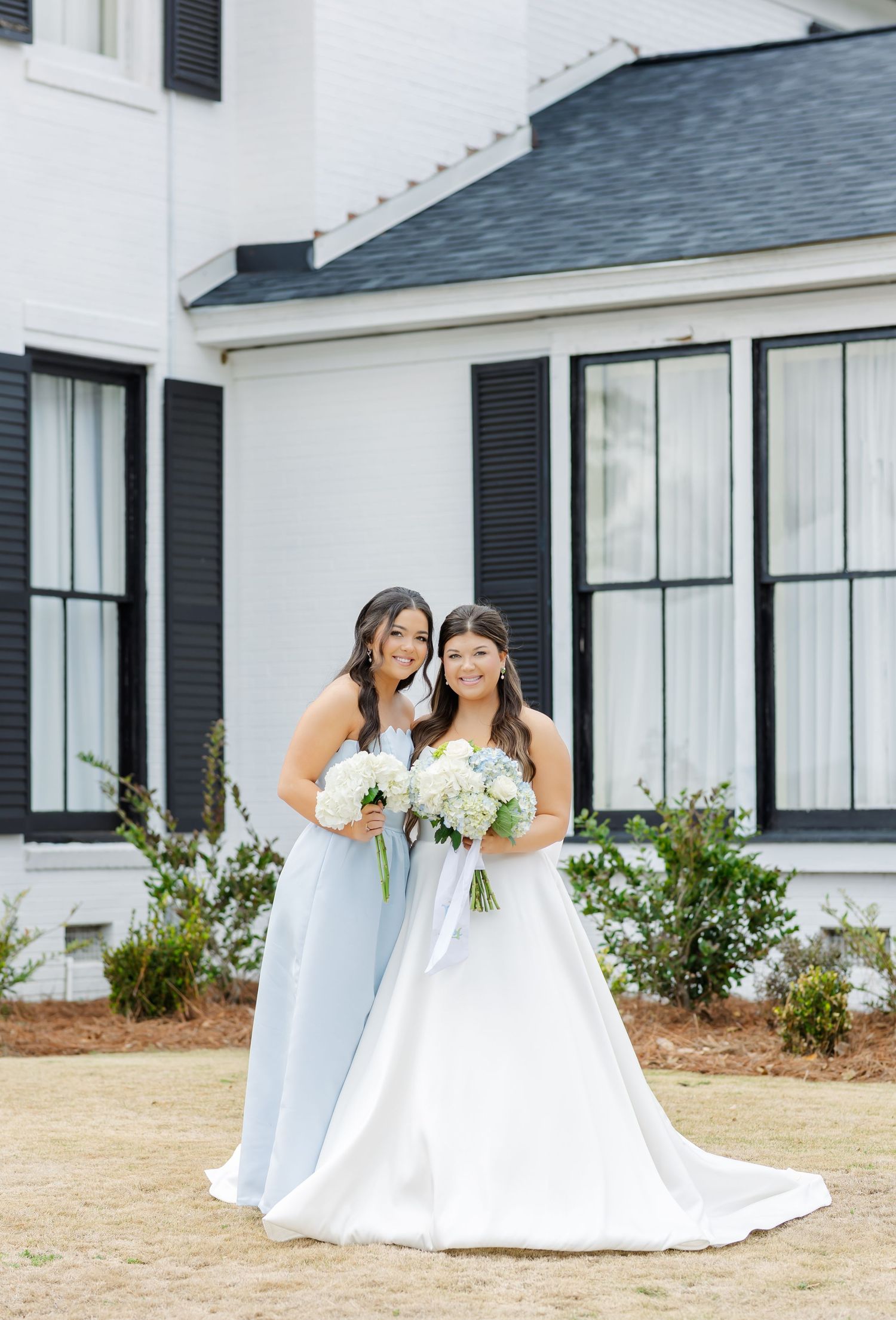 Bride in white gown and bridesmaid in light blue dress holding white bouquets outside a white house.