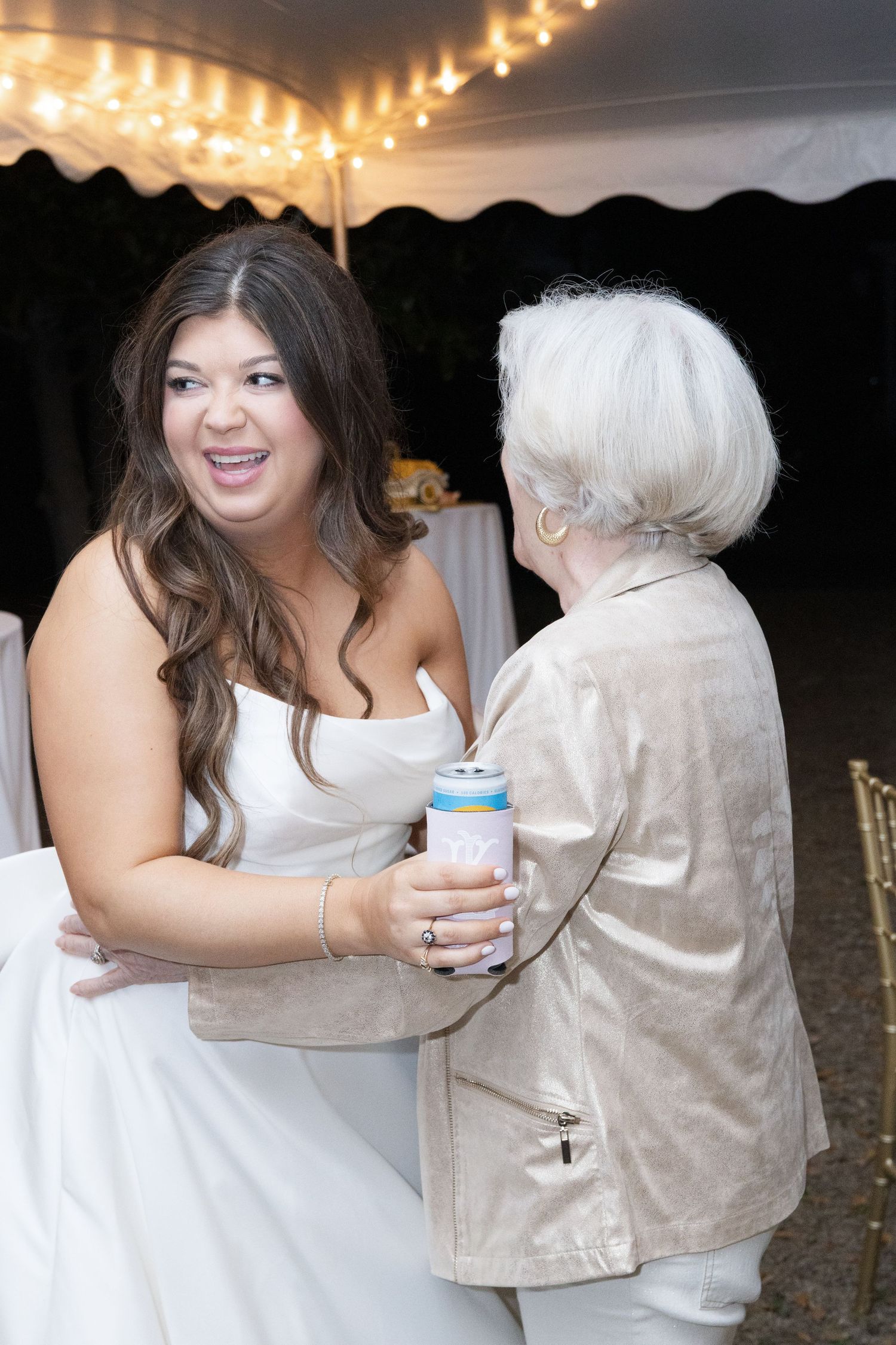 Bride in white gown laughing while dancing with elderly woman in gold jacket at outdoor reception.