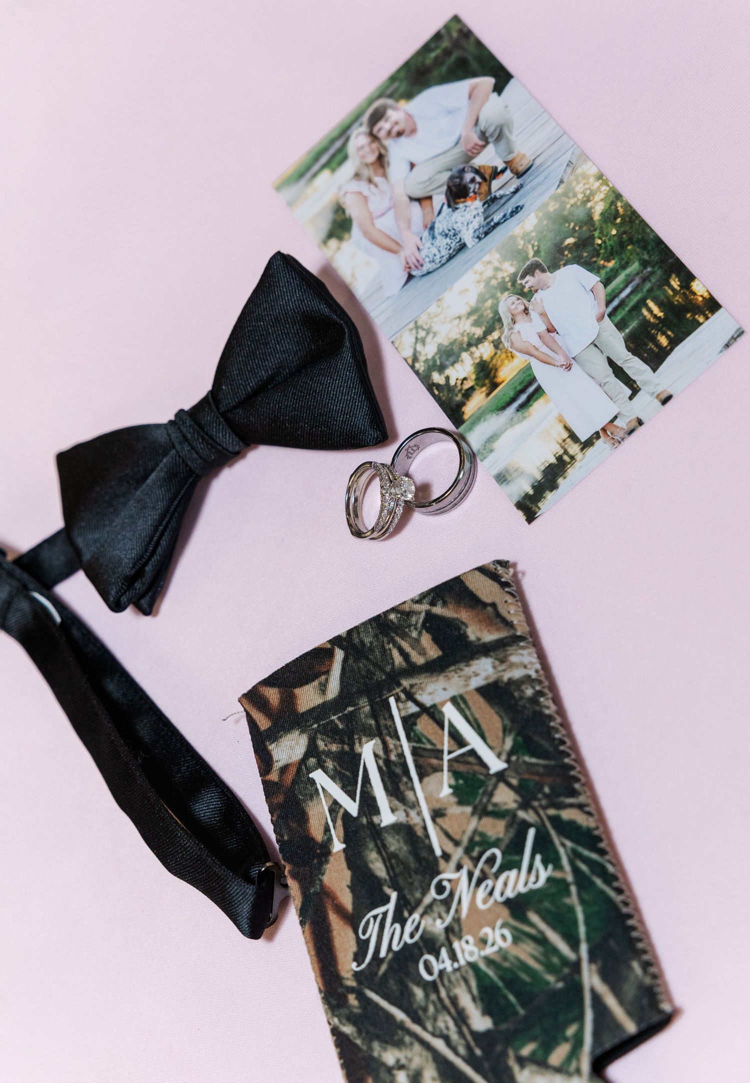 Wedding flat lay featuring black bow tie, gold rings, camo koozie with monogram M|A The Neals 04.18.26, and couple photos.