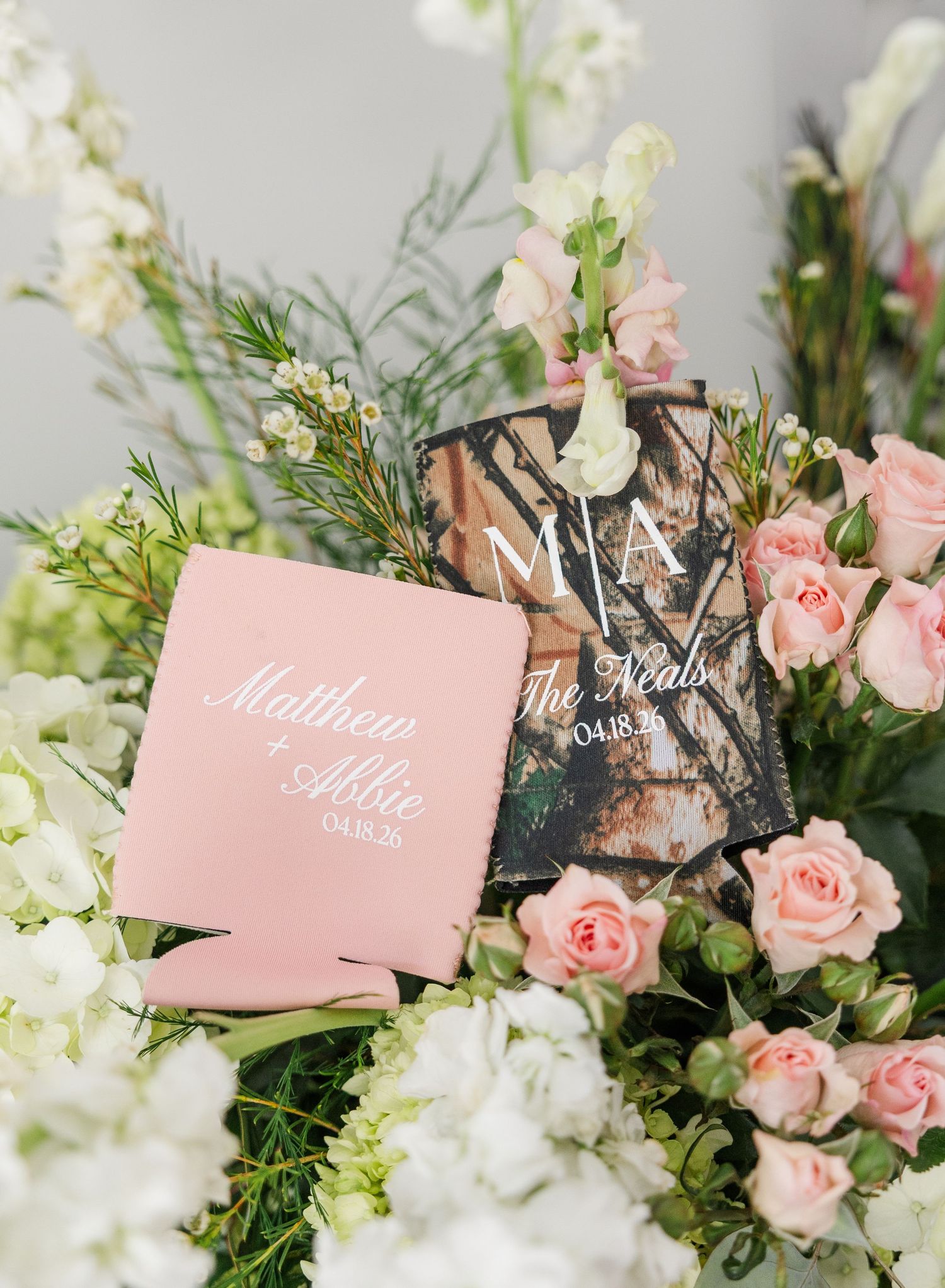 Pink and camo wedding koozies for Matthew and Abbie dated 04.18.25 displayed among pink roses and white flowers.