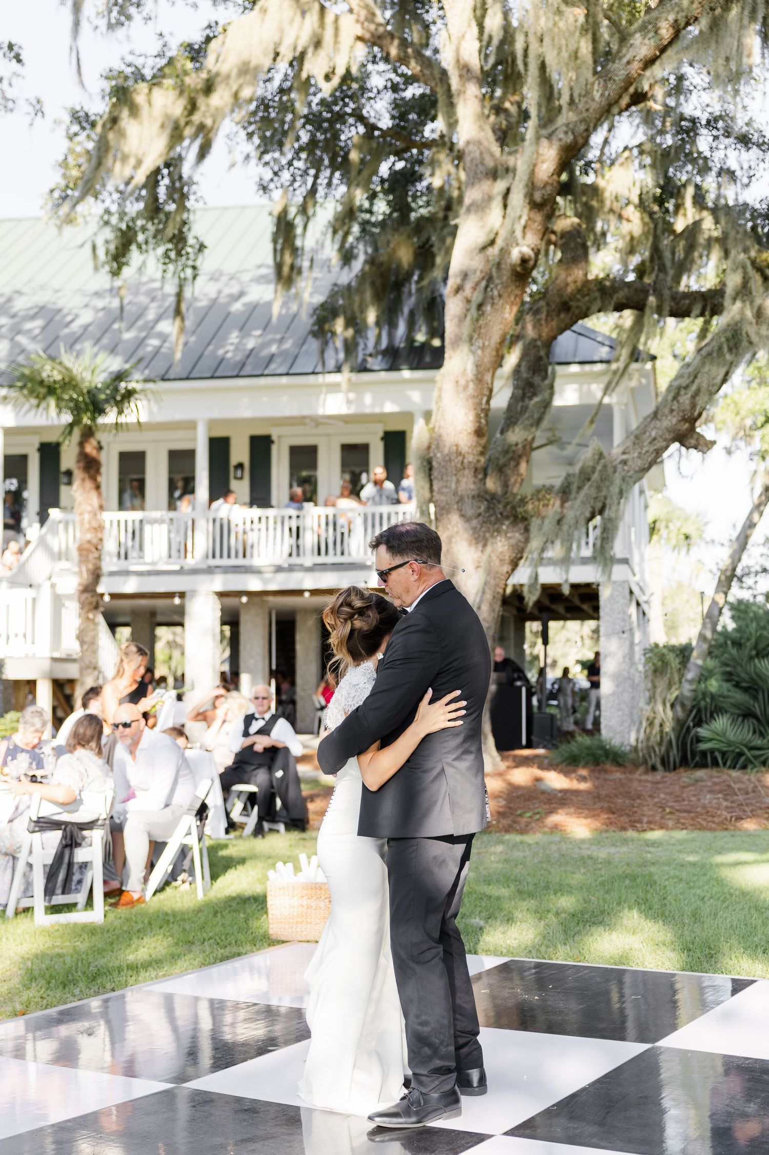 Bride and groom share first dance on checkered floor at outdoor wedding reception under Spanish moss trees.