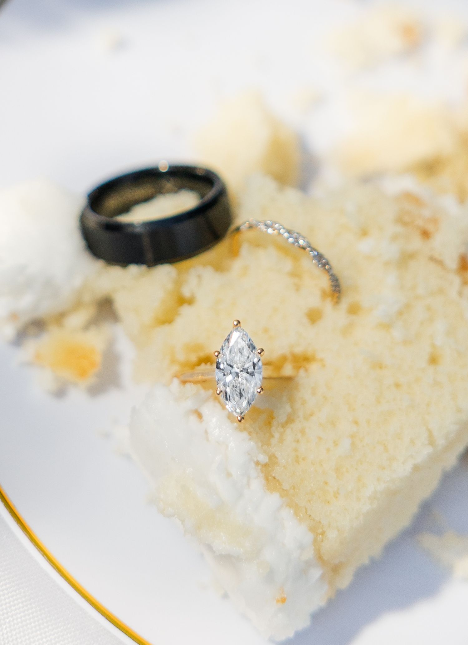 Marquise diamond engagement ring and black tungsten wedding band displayed on a slice of white wedding cake on a gold-rimmed plate.