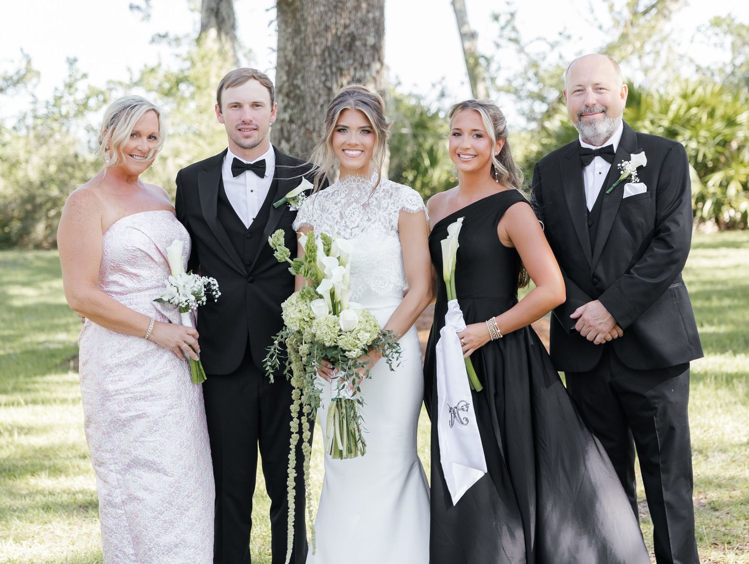 Wedding party of five posing outdoors, bride in white gown with bouquet, groom in tuxedo, guests in formal attire.