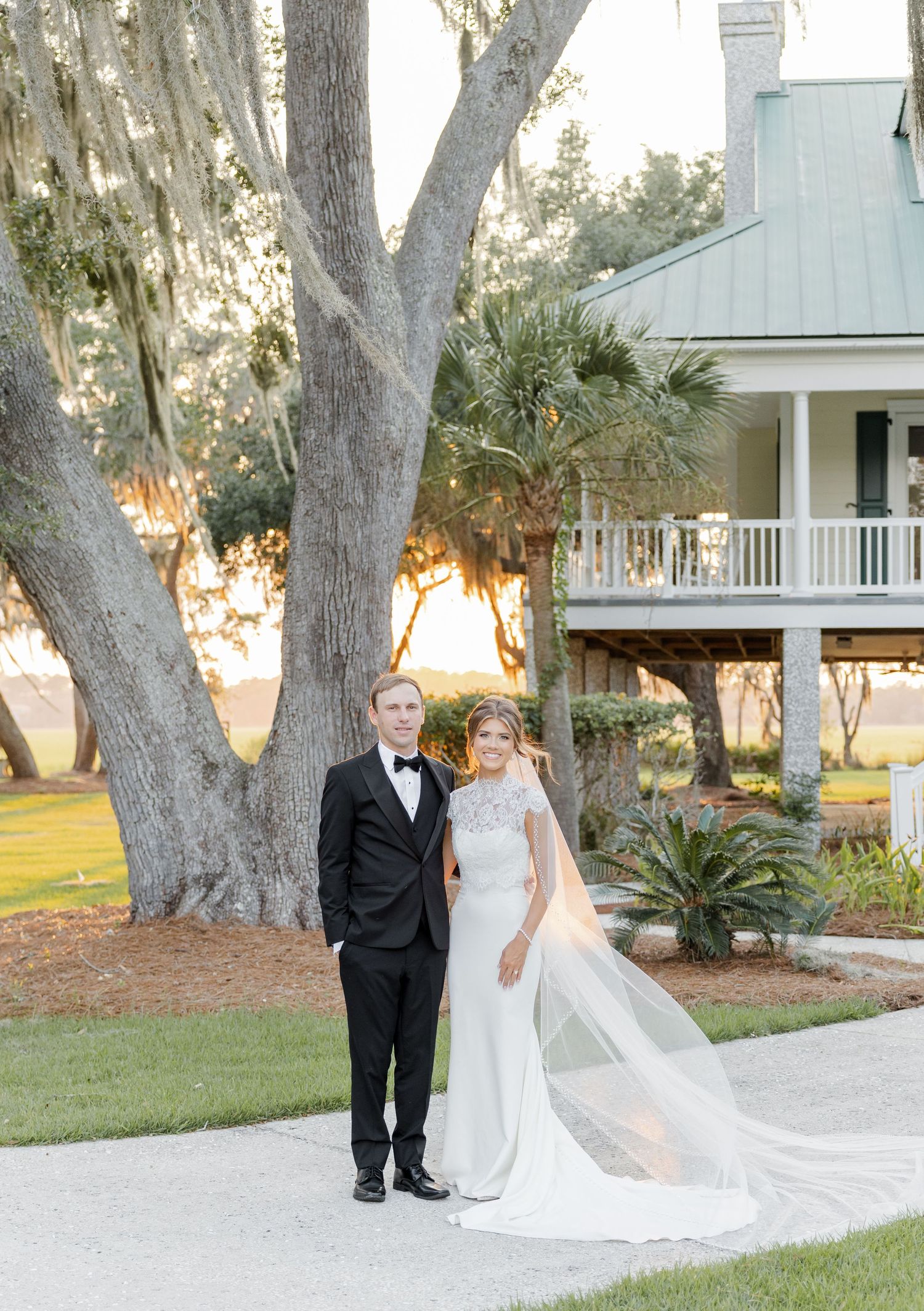 Bride in white gown with veil and groom in black tuxedo pose outside a Southern plantation home at sunset.