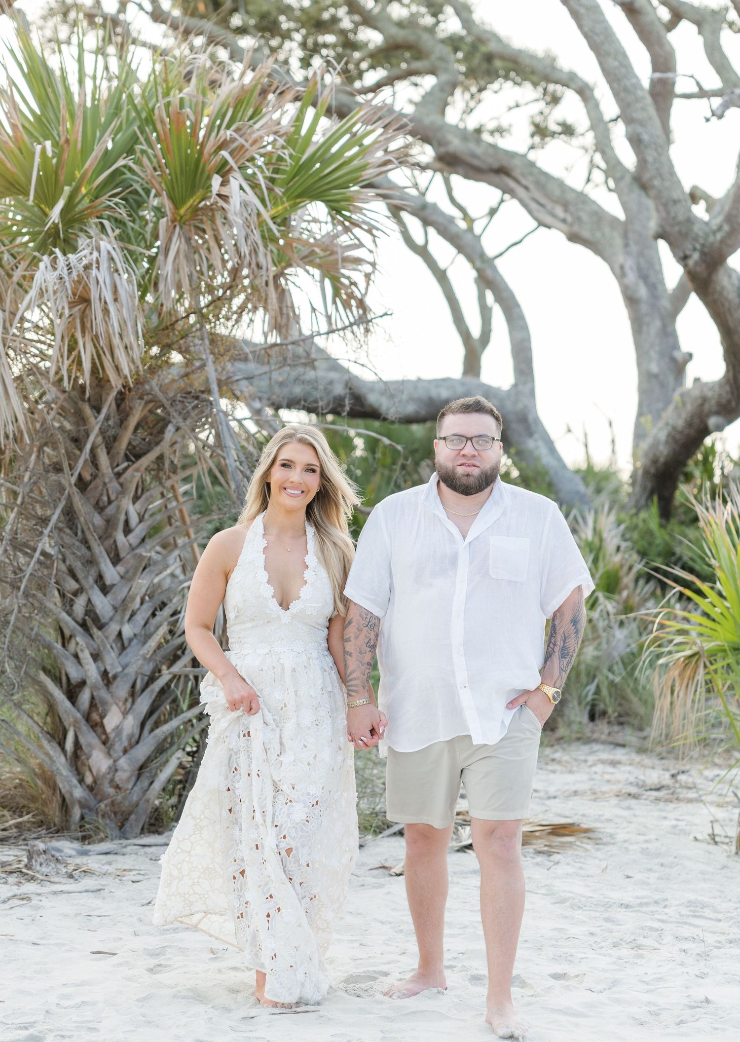 Couple in white attire walking on sandy beach with palm trees, woman in lace dress, man in white shirt and khaki shorts.