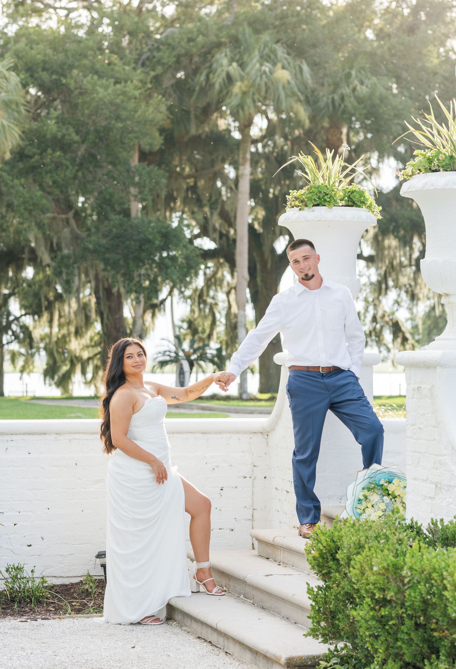 Engaged couple poses playfully on white steps holding hands surrounded by lush green trees.