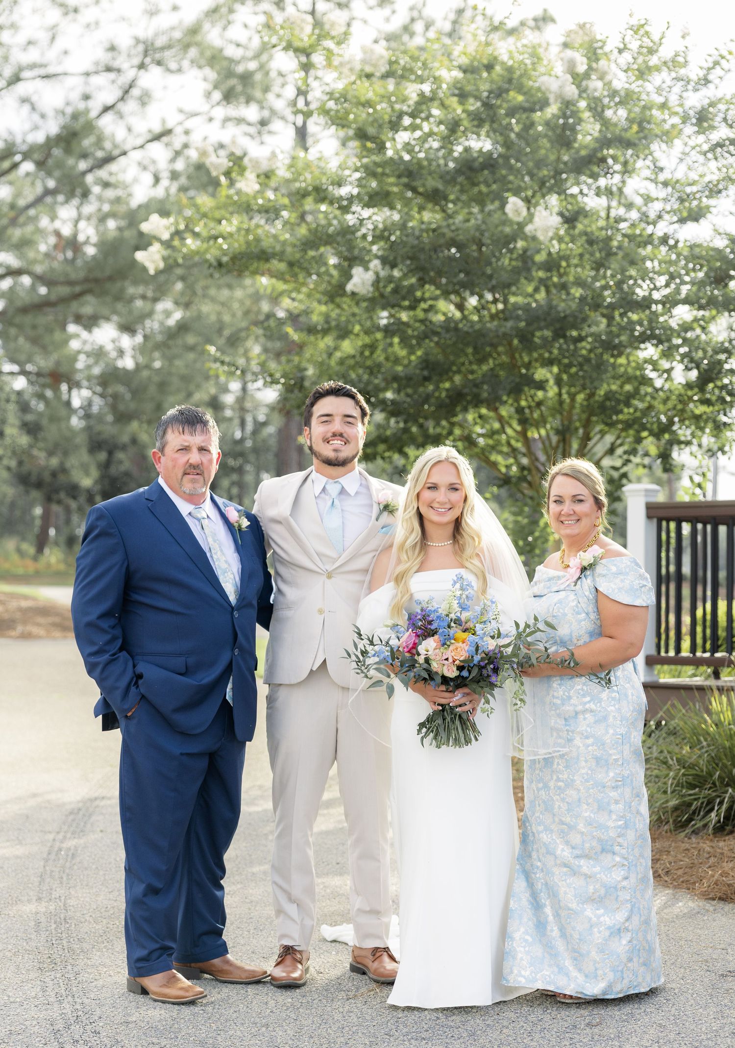 Wedding couple poses with family members outdoors, bride holding colorful bouquet in natural light.